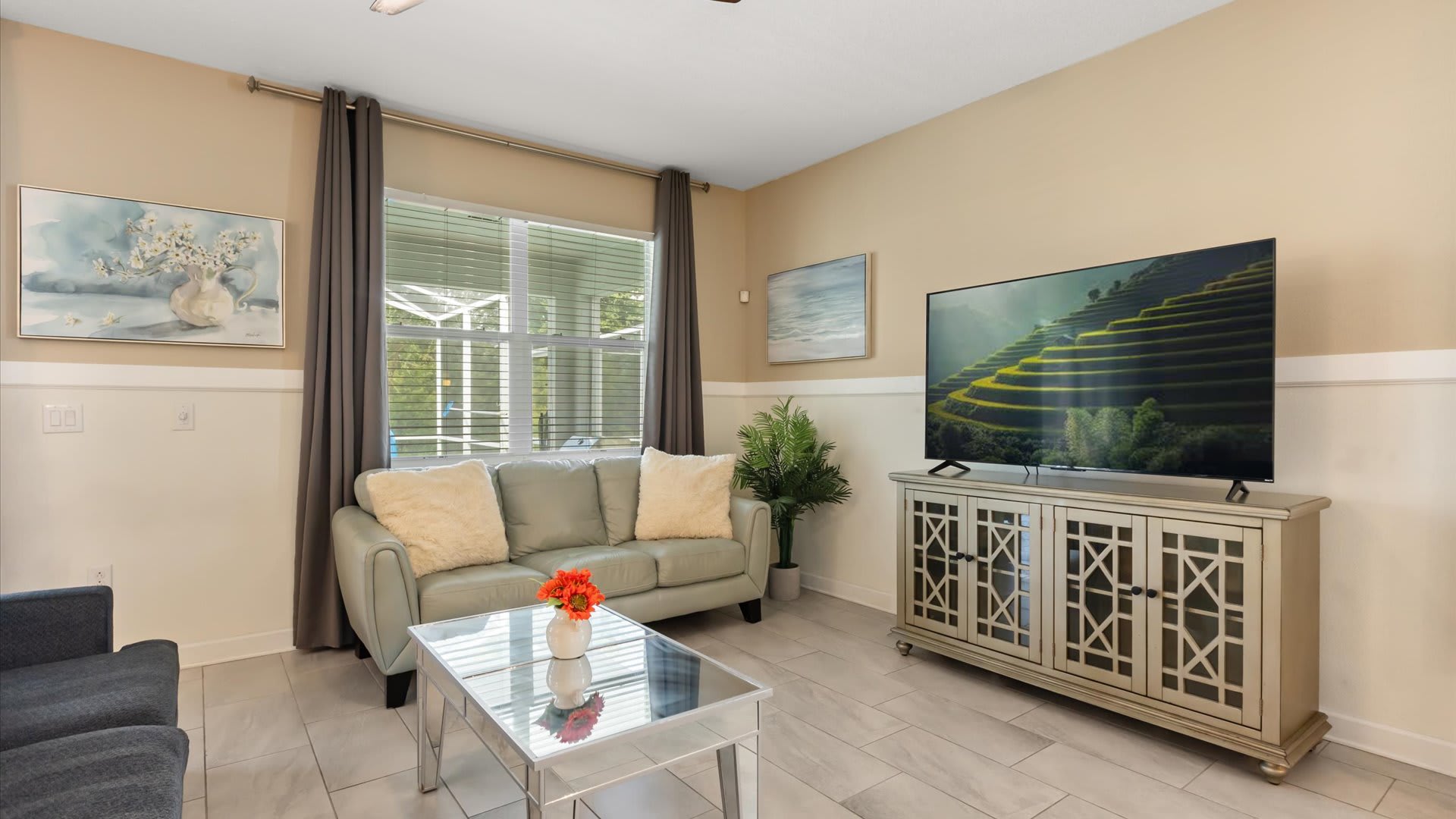 Cozy living room featuring a light green loveseat with fluffy white pillows, a large flat-screen TV on a decorative console, and a mirrored coffee table, with a window providing natural light.