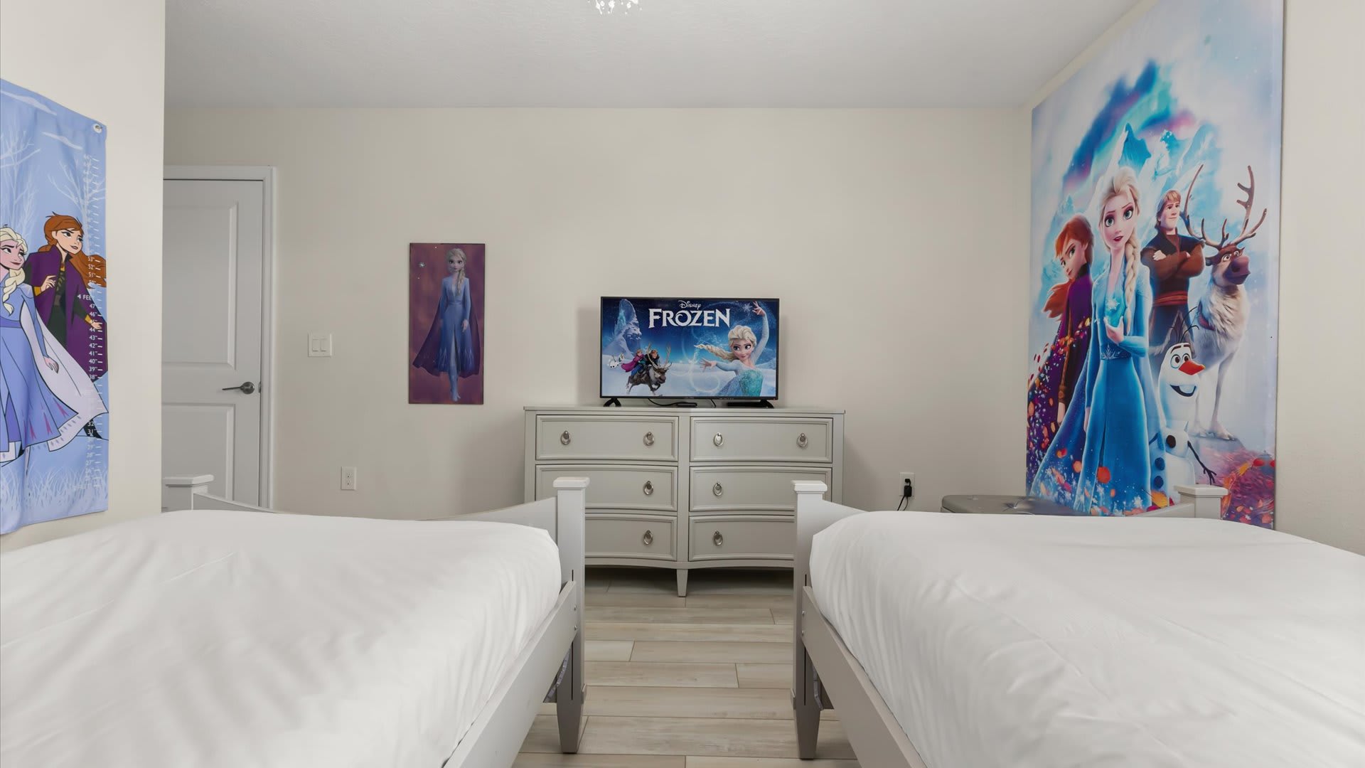 This Frozen-themed bedroom shows a white dresser with silver pulls, topped with a flat-screen TV displaying Frozen, positioned between two twin beds. A large Frozen mural covers the wall to the right.
