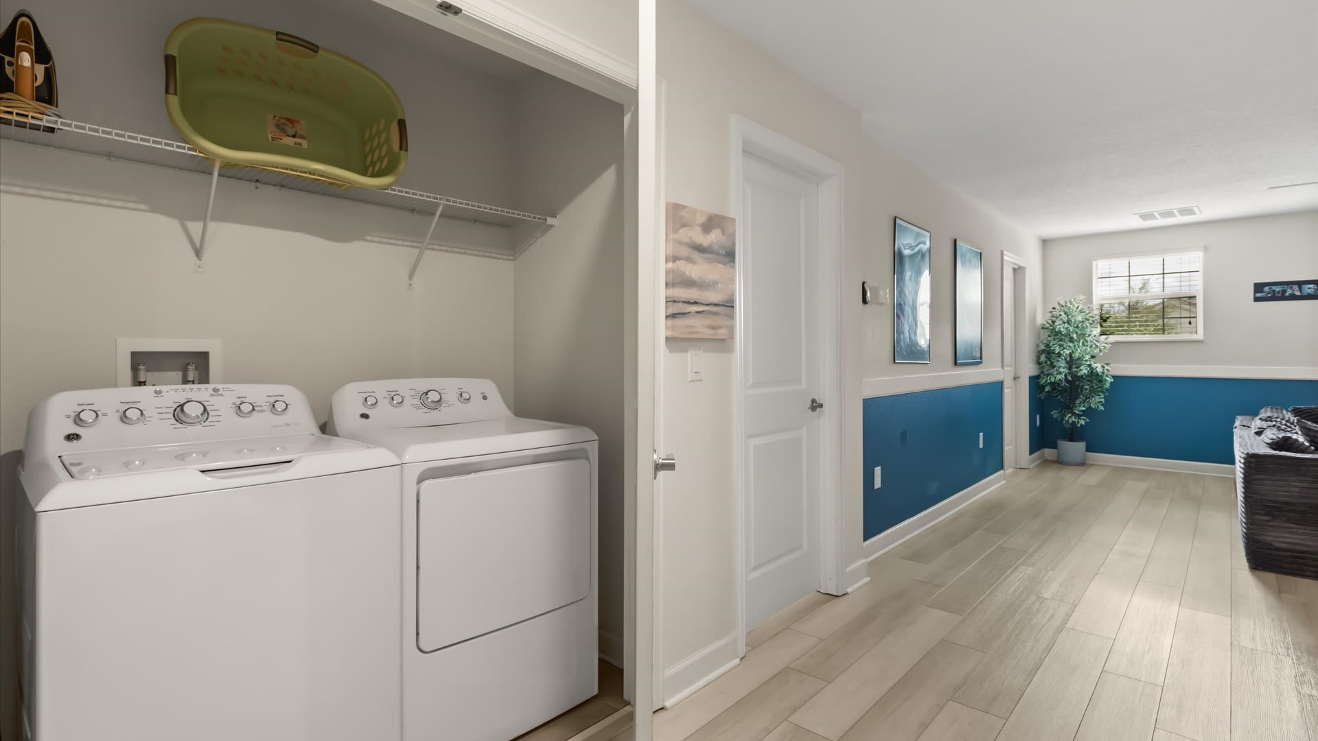 Dedicated laundry closet featuring a white top-loading washing machine and a matching dryer, with a green laundry basket stored on a wire shelf above. Adjacent is a hallway with blue wainscoting and light wood-look flooring.