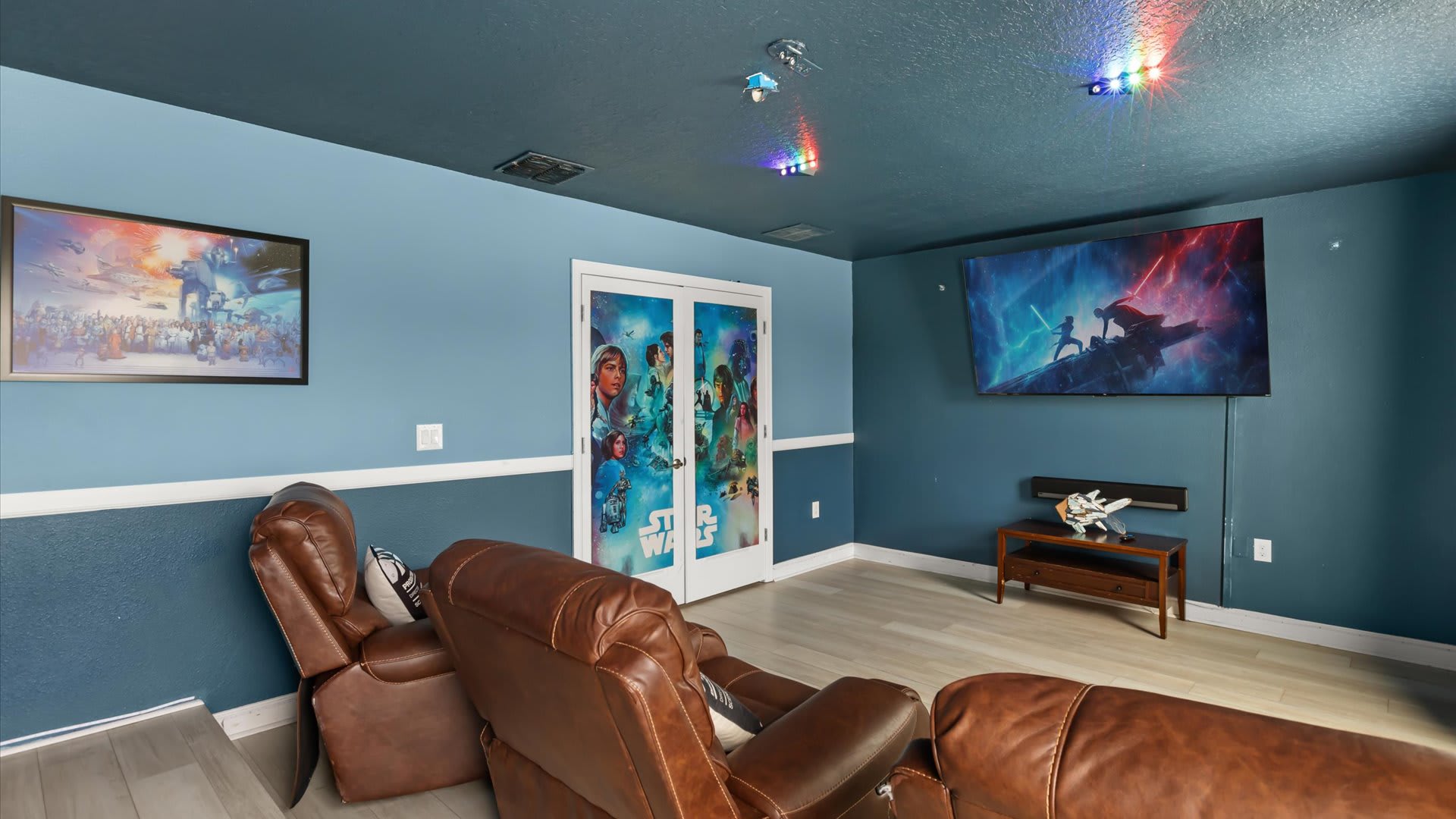 A Star Wars-themed media room features two rows of brown leather recliners facing a large wall-mounted TV, with blue and white walls and a dark blue ceiling adorned with colorful LED lights. French doors with Star Wars decals are visible.