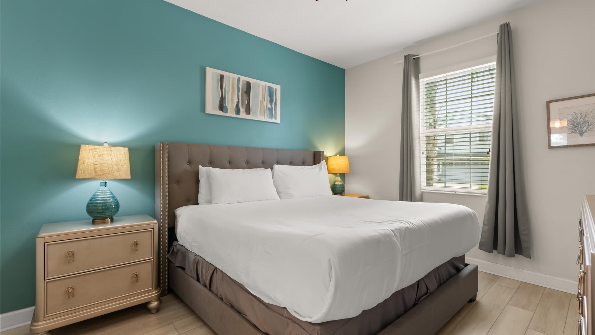 Inviting bedroom with a king-sized bed featuring a gray tufted headboard, flanked by two light-colored nightstands with vibrant blue lamps. A teal accent wall behind the bed adds a pop of color, complemented by light wood-look flooring.