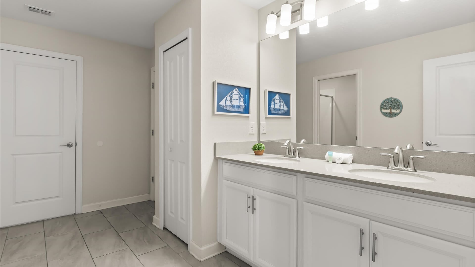 Modern bathroom featuring a double vanity with light gray quartz countertops, white shaker-style cabinets, and two chrome faucets. Nautical sailboat artworks adorn the light-colored walls above the vanity.