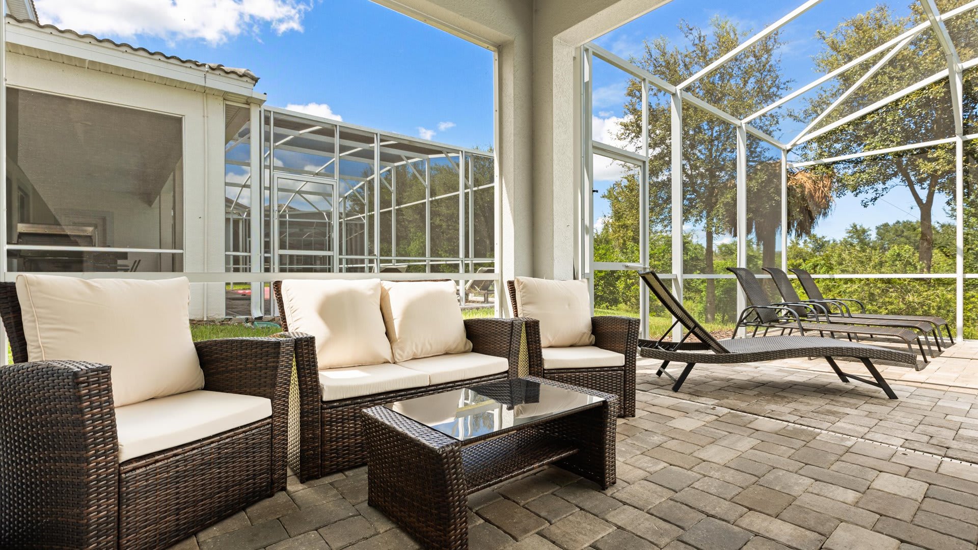 A covered outdoor patio area features a dark brown wicker sofa and two armchairs with cream cushions, centered around a glass-top coffee table. Several lounge chairs are visible on the open paver deck next to the screened enclosure.