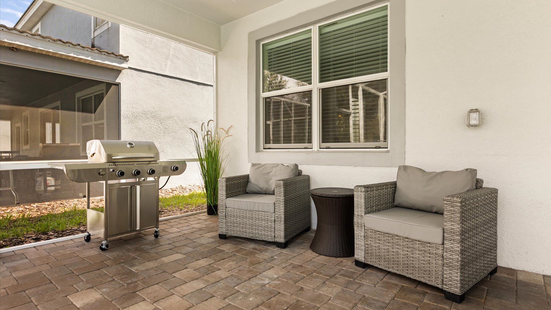 A covered outdoor patio area features two gray wicker armchairs with cushions and a small side table, positioned next to a stainless steel BBQ grill. A large window of the house is visible in the background.