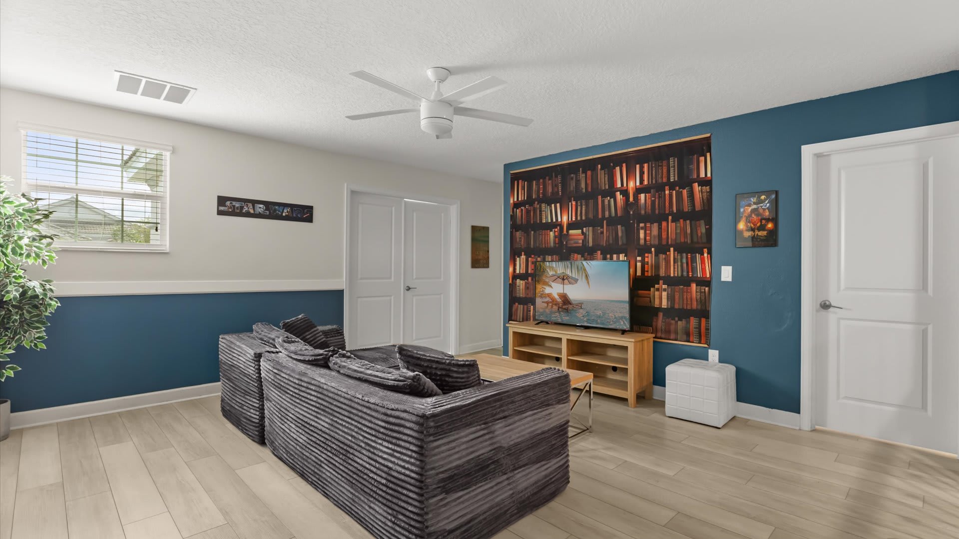A casual lounge area features a gray sectional sofa facing a TV mounted against a wall with a bookshelf-themed wallpaper, light wood-look flooring, and a white ceiling fan. The walls are two-toned blue and white.