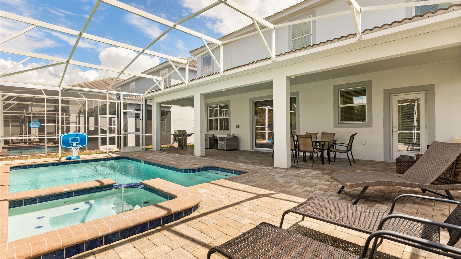 A spacious screened-in patio area features a rectangular swimming pool with an adjacent hot tub, surrounded by paver decking. A covered section includes an outdoor dining table and lounge chairs, with the house exterior visible.