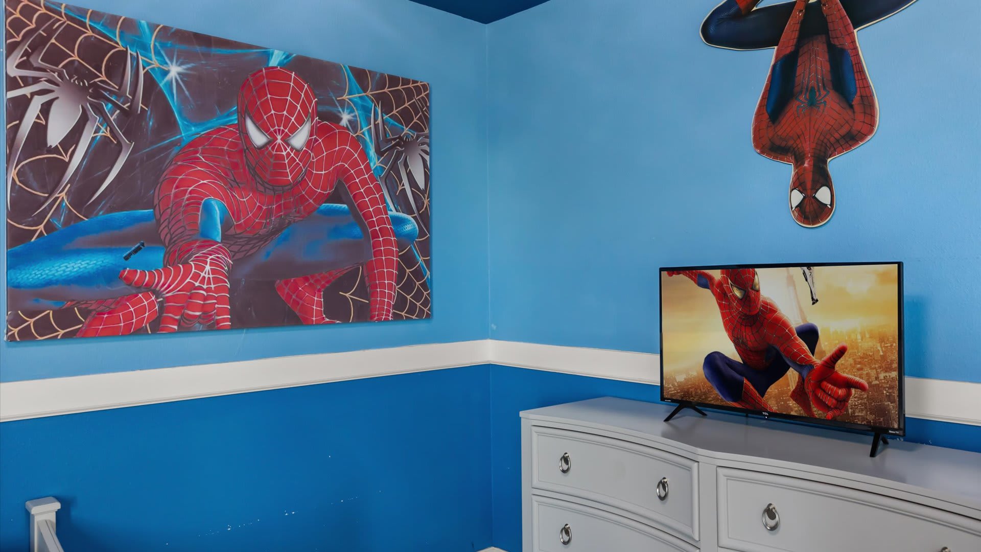 A close-up view in the Spiderman-themed bedroom highlights a large canvas art piece featuring Spiderman in action, positioned above a white dresser with a flat-screen TV displaying a Spiderman scene. An upside-down Spiderman decal is on the wall.
