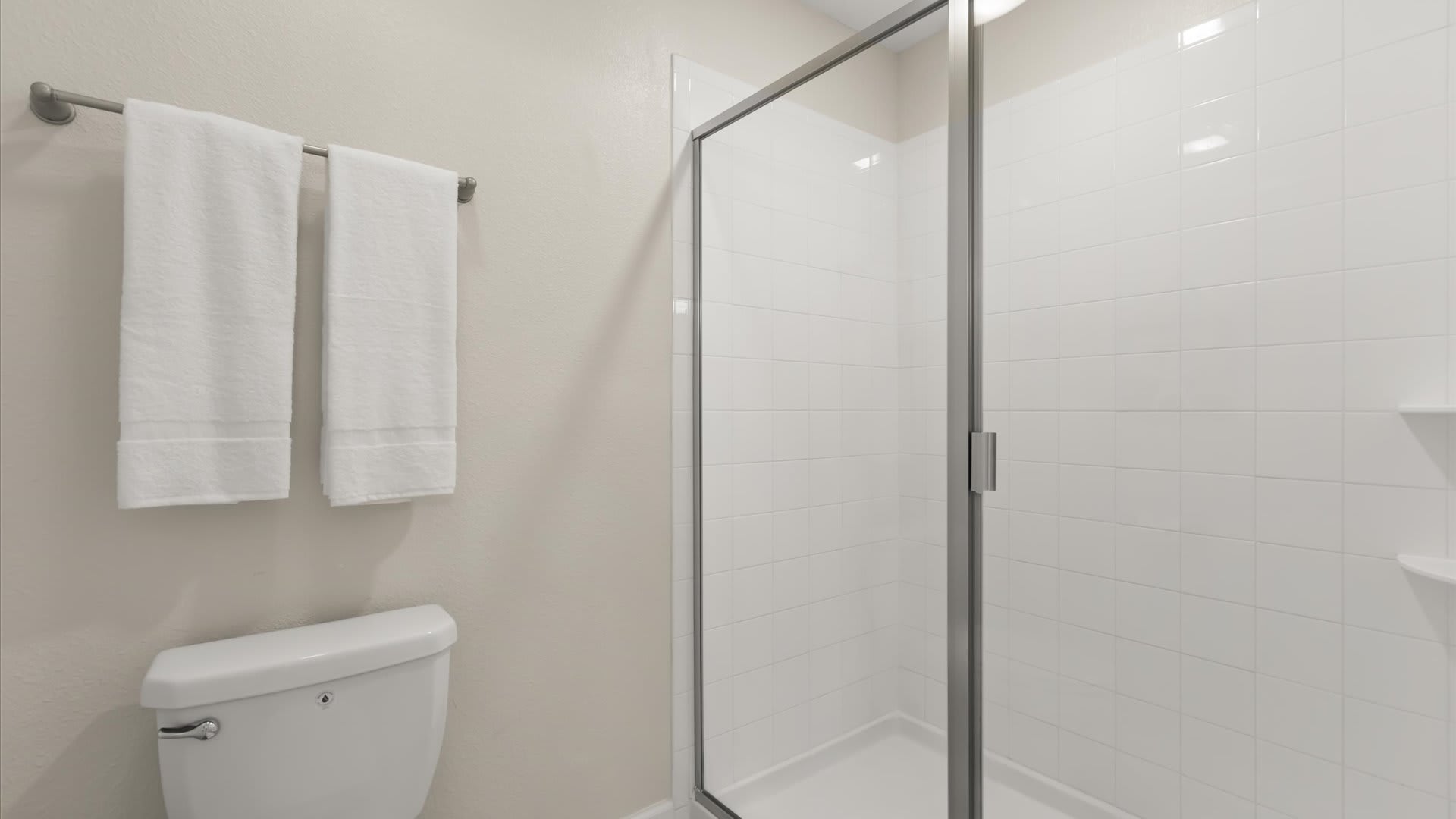 Bright bathroom showcasing a white toilet and a glass-enclosed walk-in shower with white subway tiles and a chrome shower head. Two neatly folded white towels hang on a wall-mounted bar.