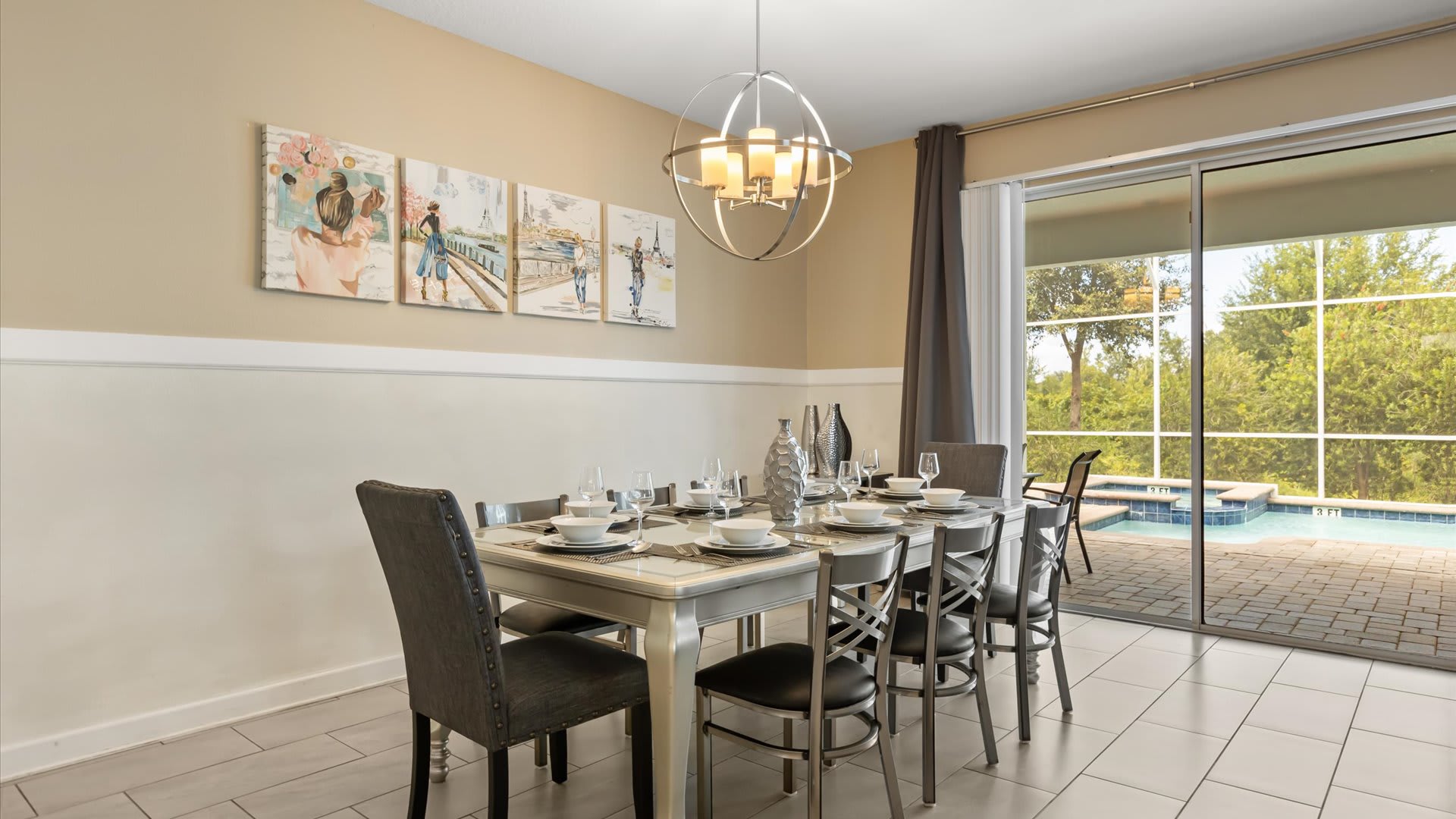 Elegant dining room with a long rectangular table set for eight, featuring dark upholstered chairs and a modern spherical chandelier, with sliding glass doors opening to a private pool area.