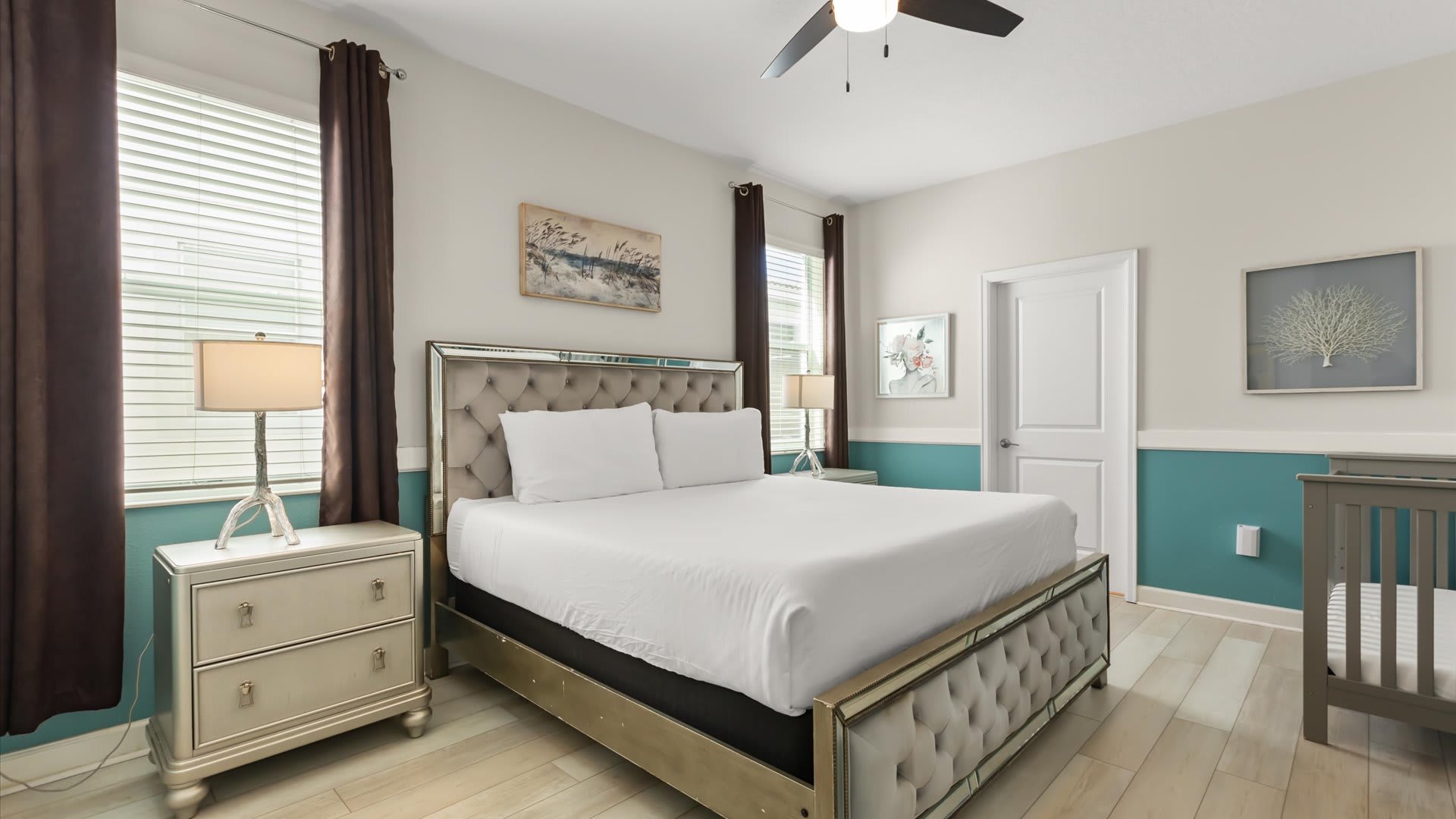 Spacious bedroom featuring a king-sized bed with a mirrored, tufted headboard, two elegant nightstands with lamps, and a baby crib. The walls are painted with a two-tone design, featuring a teal wainscoting and light beige upper section.