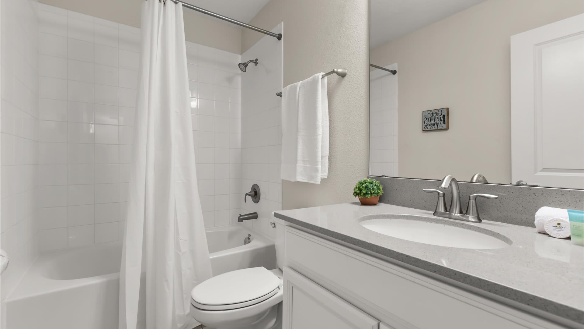 A clean bathroom features a single vanity with a light gray countertop and a white sink, next to a toilet and a white tiled tub/shower combo with a white curtain. A small green plant adds a touch of color to the vanity.