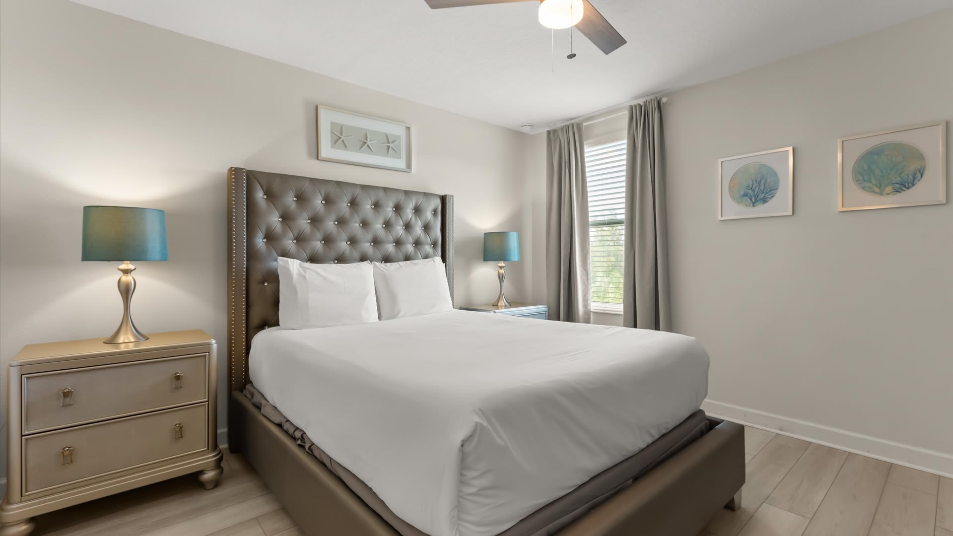 A cozy bedroom features a queen-sized bed with a tall, tufted dark gray headboard and white bedding, flanked by two nightstands with teal lamps. Coastal-themed artwork adorns the light-colored walls.