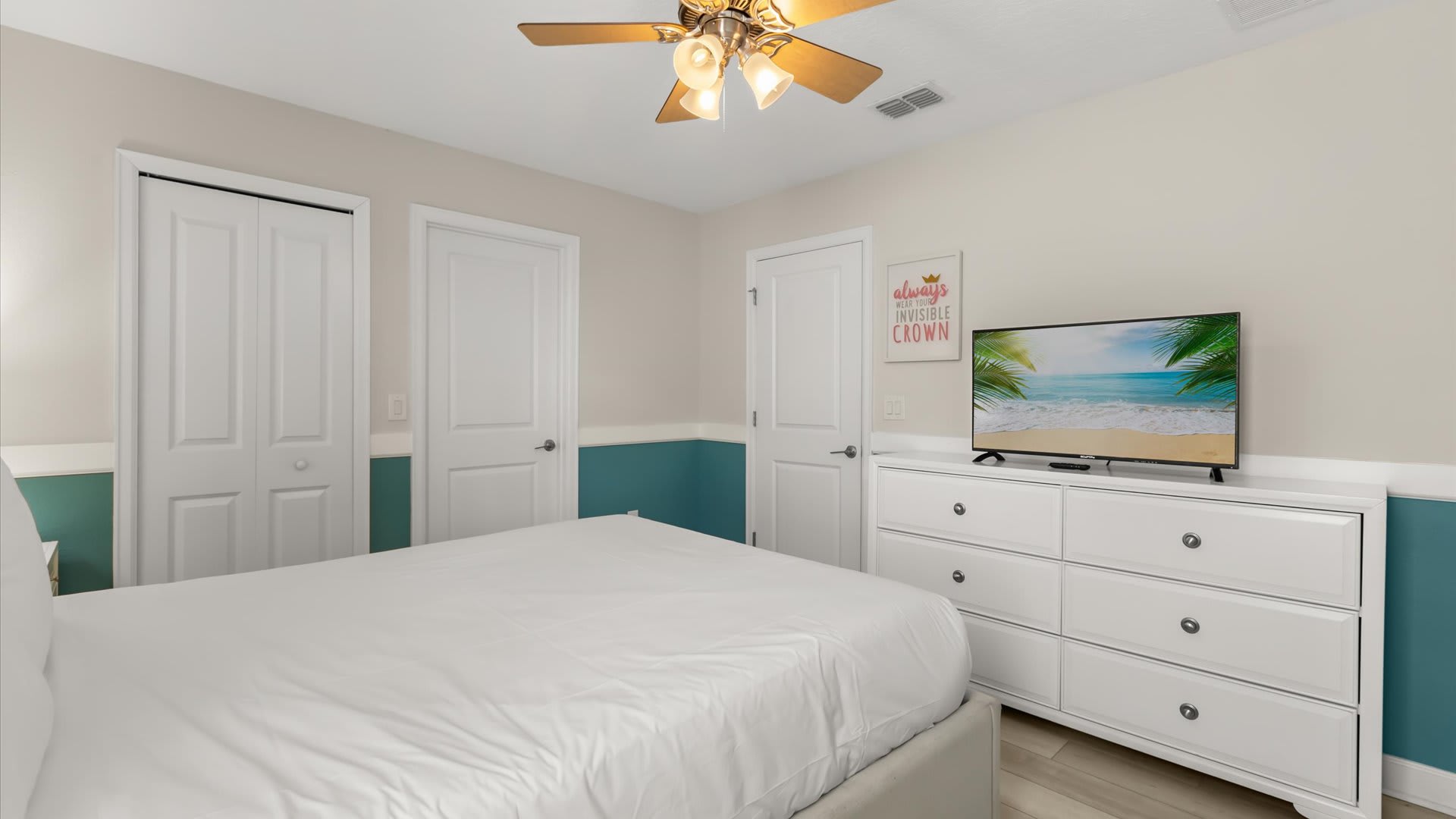 Another angle of the vibrant bedroom, showcasing a white dresser with a flat-screen TV, bifold closet doors, and a motivational wall art piece. The teal wainscoting continues around the room, adding a playful touch.
