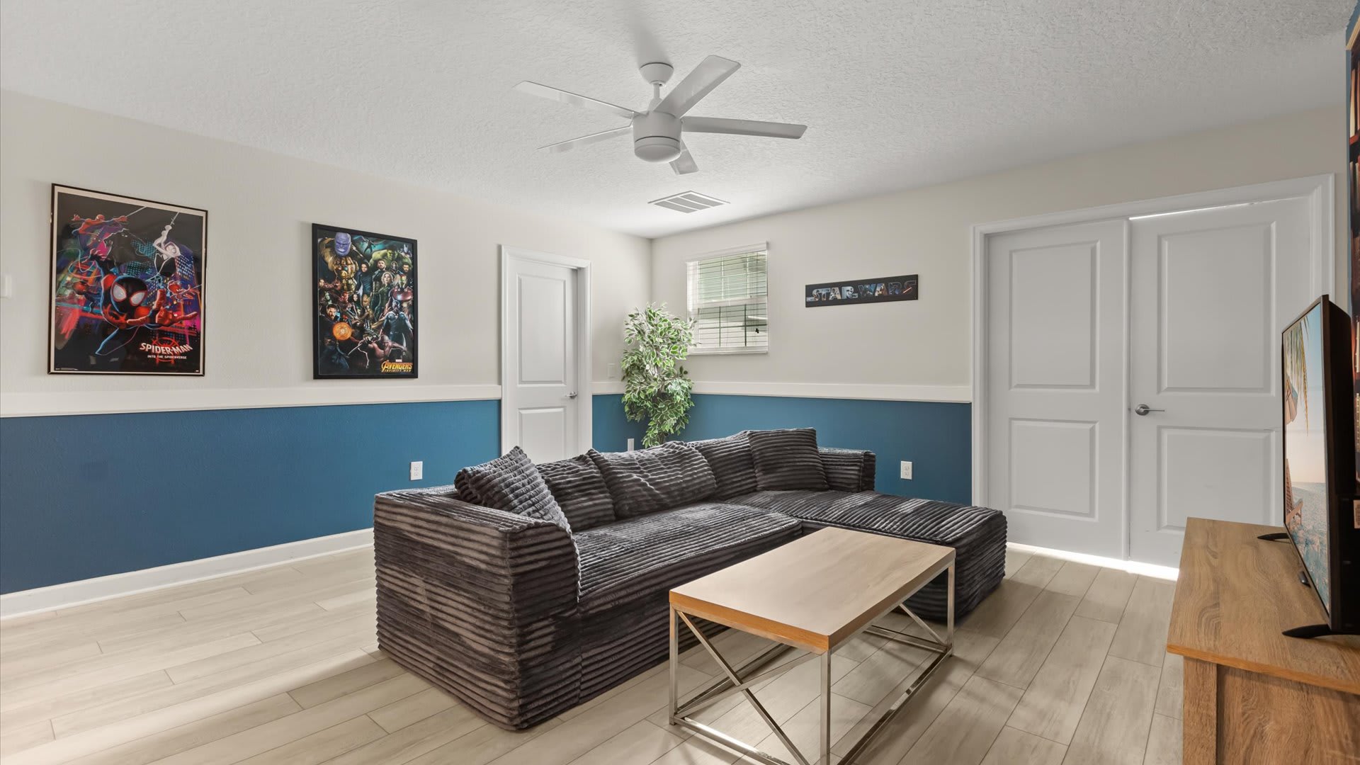 Casual game room or secondary living area with a large grey sectional sofa, a light wood coffee table, and a flat-screen TV on a matching console, decorated with superhero posters.