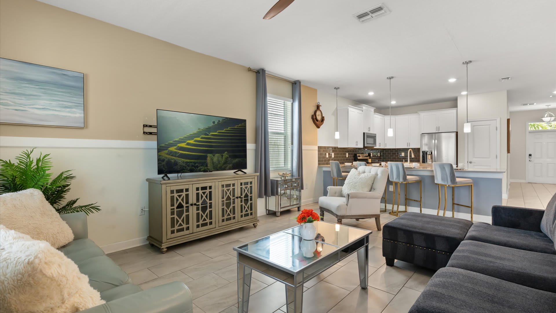 Bright open-concept living area with a large flat-screen TV on a light wood console, a grey sectional sofa, a light green loveseat, and a mirrored coffee table, leading into a modern kitchen.