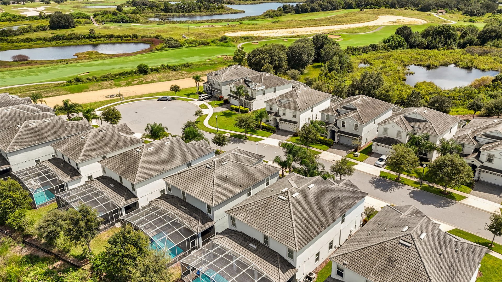 Aerial view of a residential community featuring numerous houses with screened-in pools, surrounded by lush green golf course fairways and several small lakes under a clear sky.