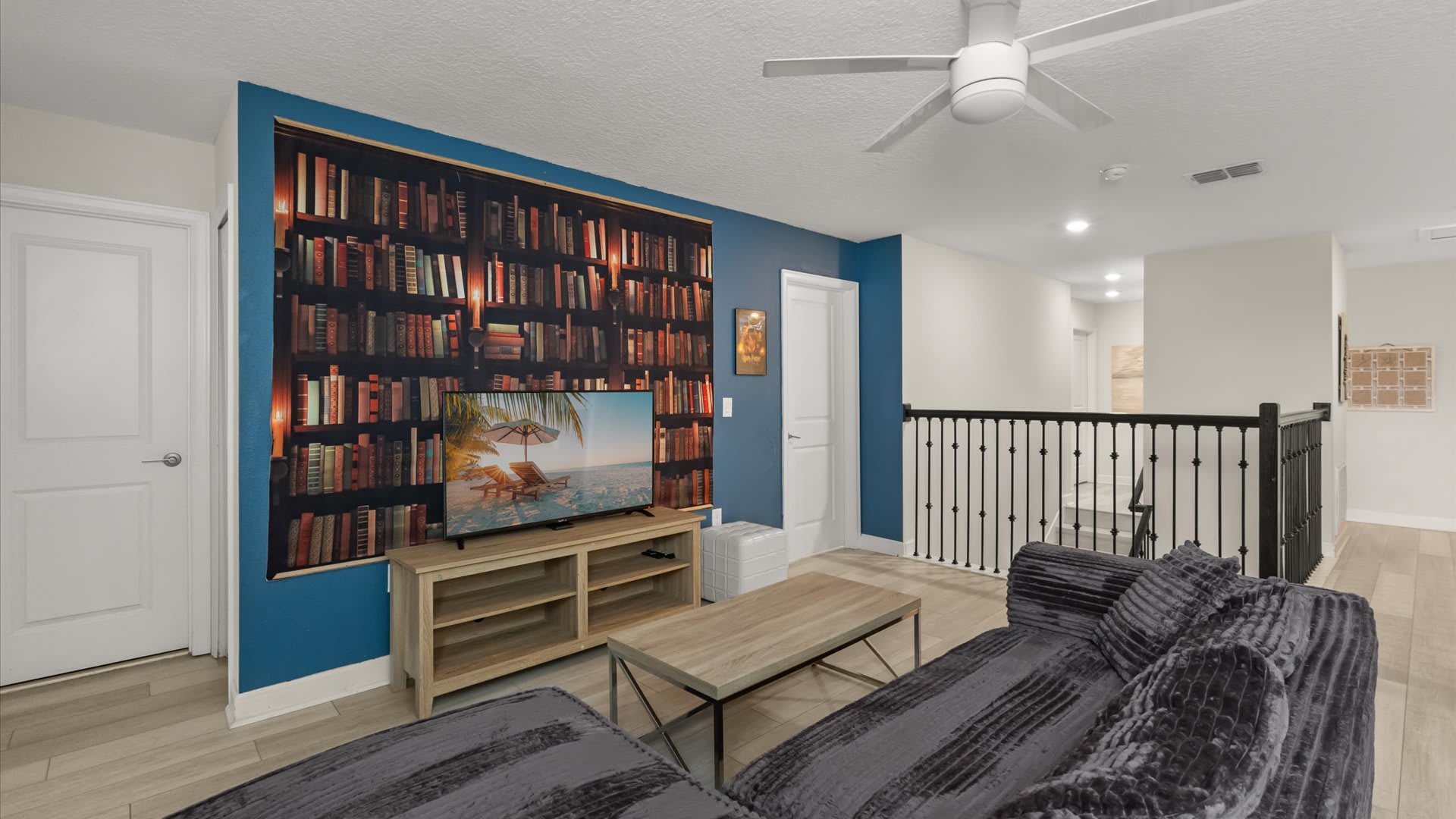 Upstairs loft area serving as a secondary living space, featuring a large grey sectional sofa, a wooden coffee table, a flat-screen TV against a bookshelf mural, and an open railing overlooking the floor below.