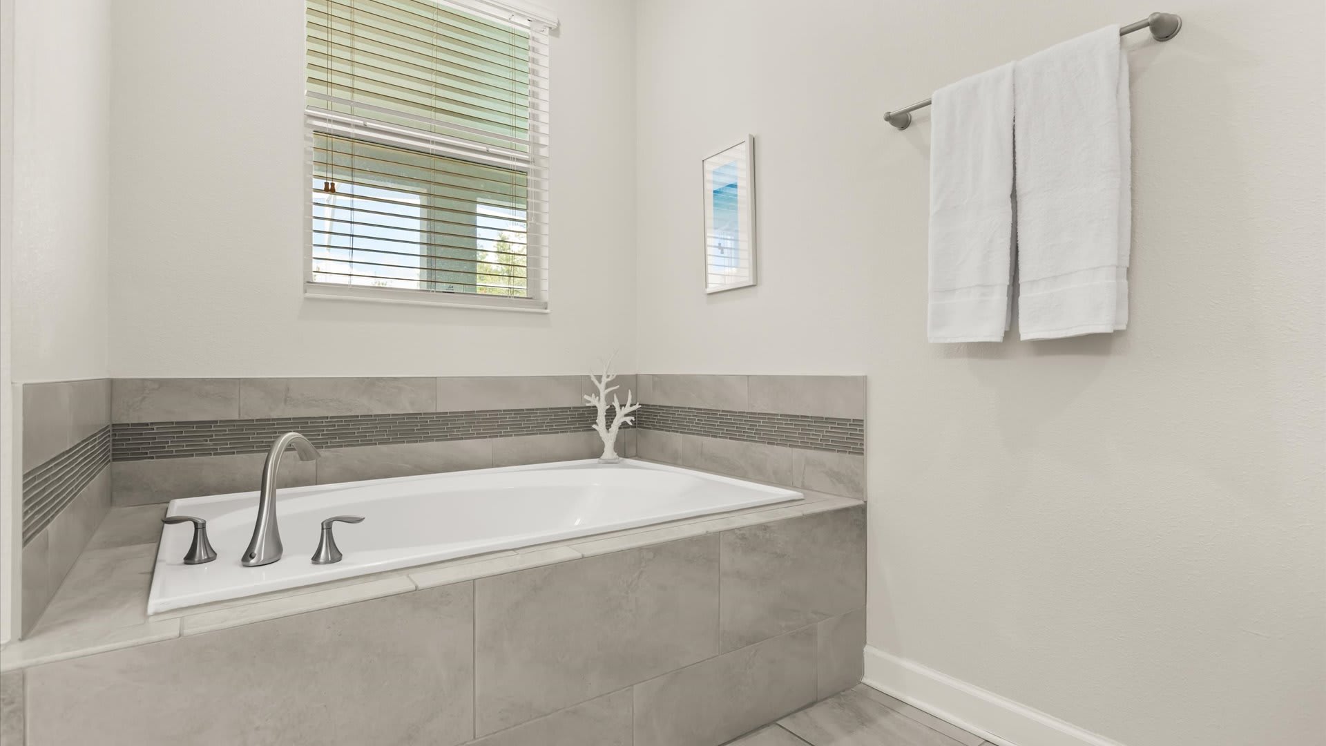 Built-in soaking tub with gray tile surround, positioned beneath a window with blinds. A decorative white coral piece and framed artwork add a touch of coastal elegance to the space.
