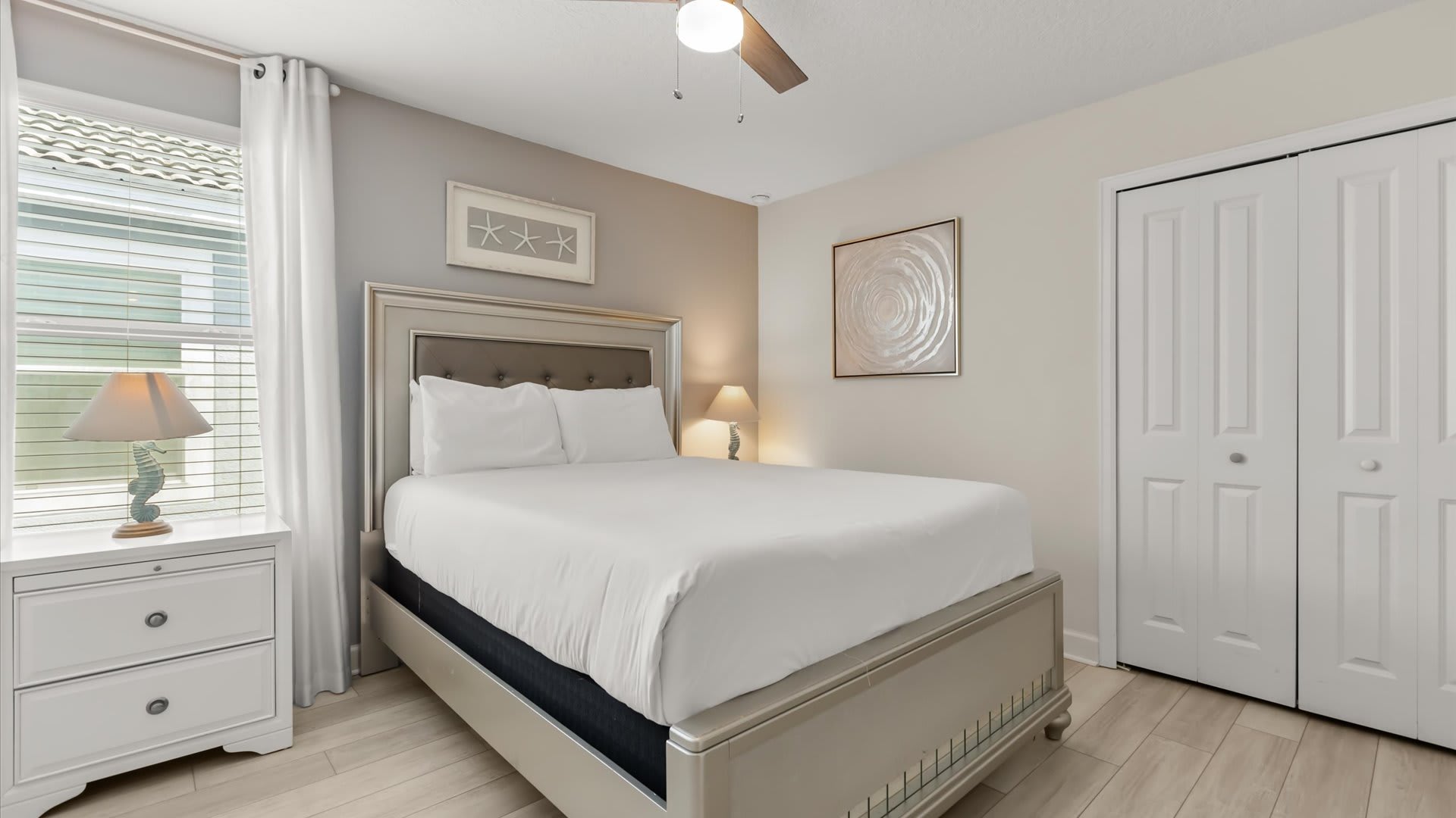 Serene bedroom featuring a silver-framed queen bed with a tufted headboard and white bedding, complemented by a white nightstand with a seahorse-themed lamp. Coastal starfish artwork hangs above the bed, and a ceiling fan provides comfort.