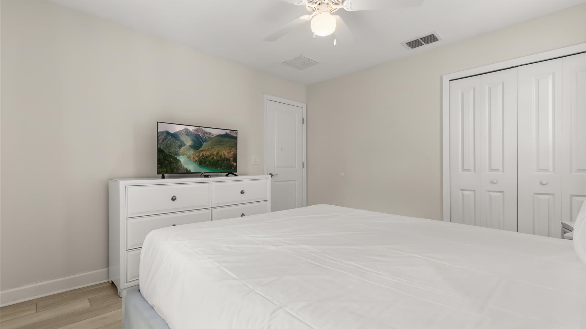Another perspective of the bedroom, showing a flat-screen TV on a white dresser and a closet with bi-fold doors. The room maintains a neutral color palette with light beige walls and light wood-look flooring.