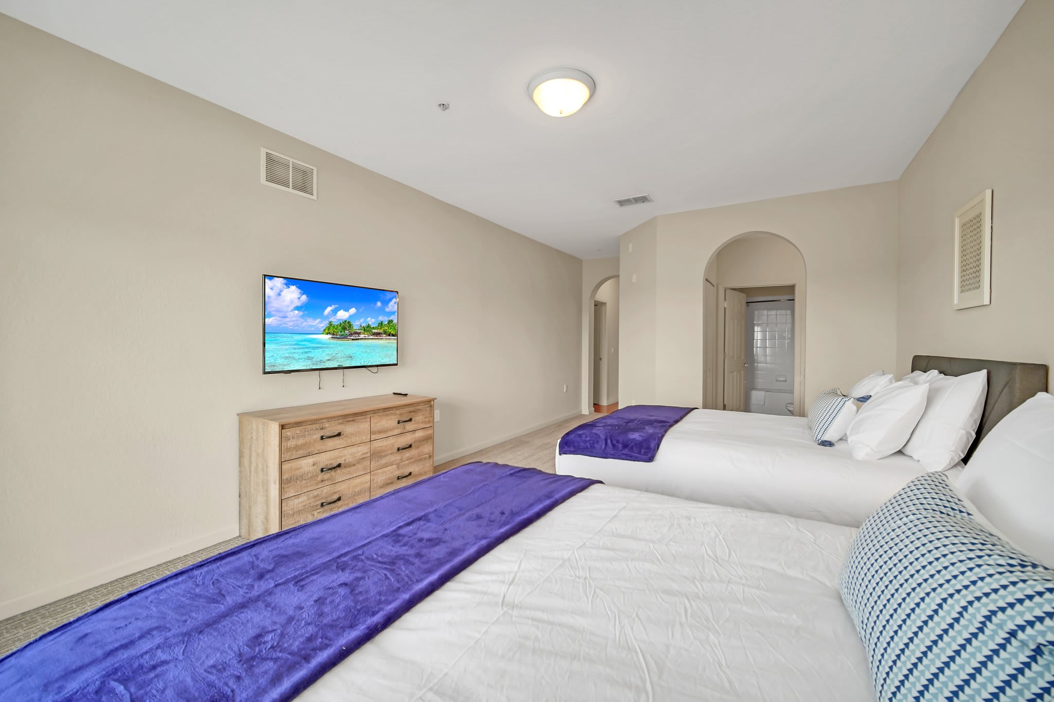 View of the second bedroom showcasing two queen beds with white linens and purple throws, a wall-mounted TV, and an archway leading to an adjacent room, all on a light patterned carpet.