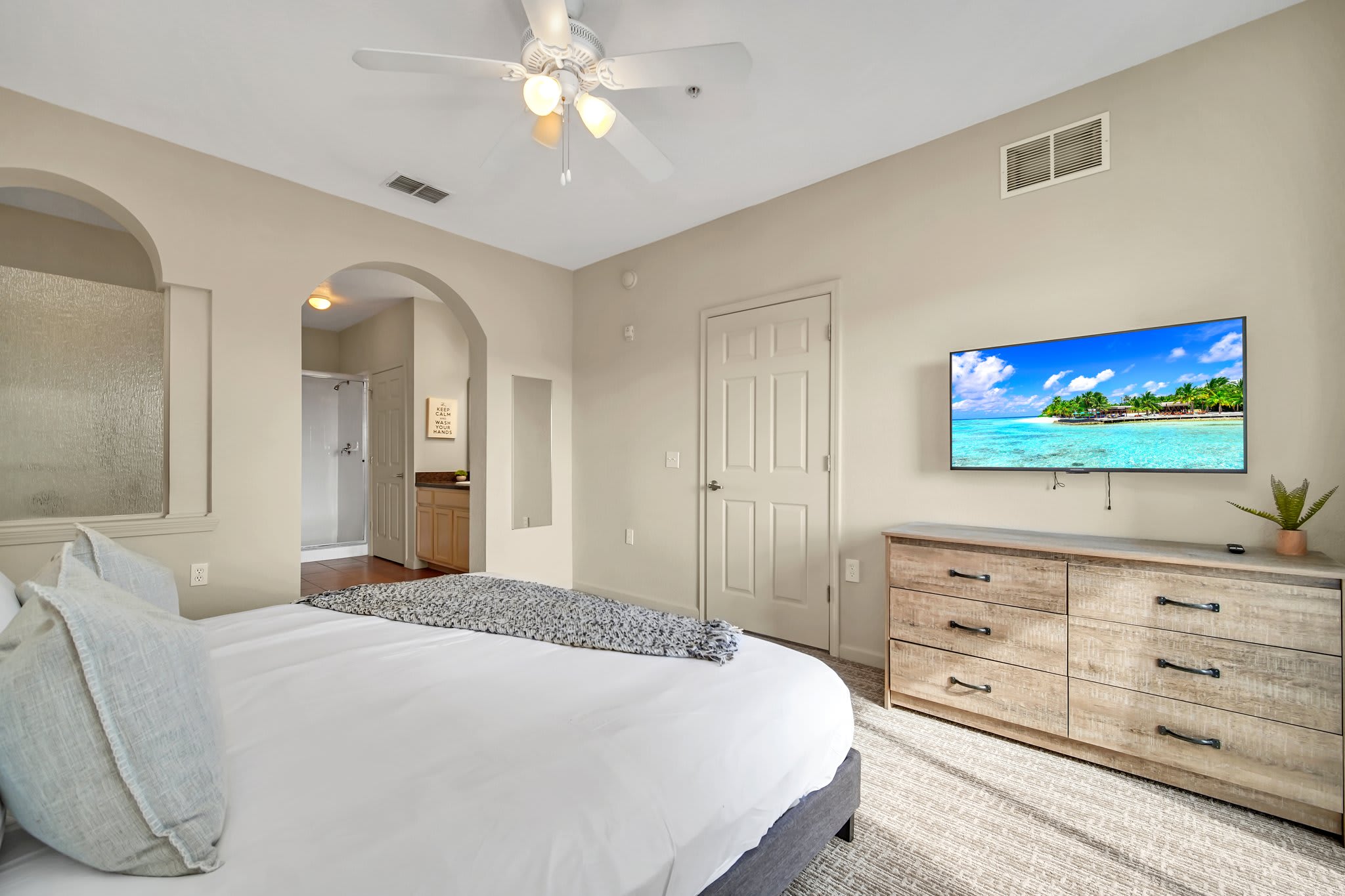 A cozy bedroom with a large bed featuring a white duvet and grey throw, a light wood dresser with a mounted flat-screen TV, and an arched entryway leading to an en-suite bathroom.