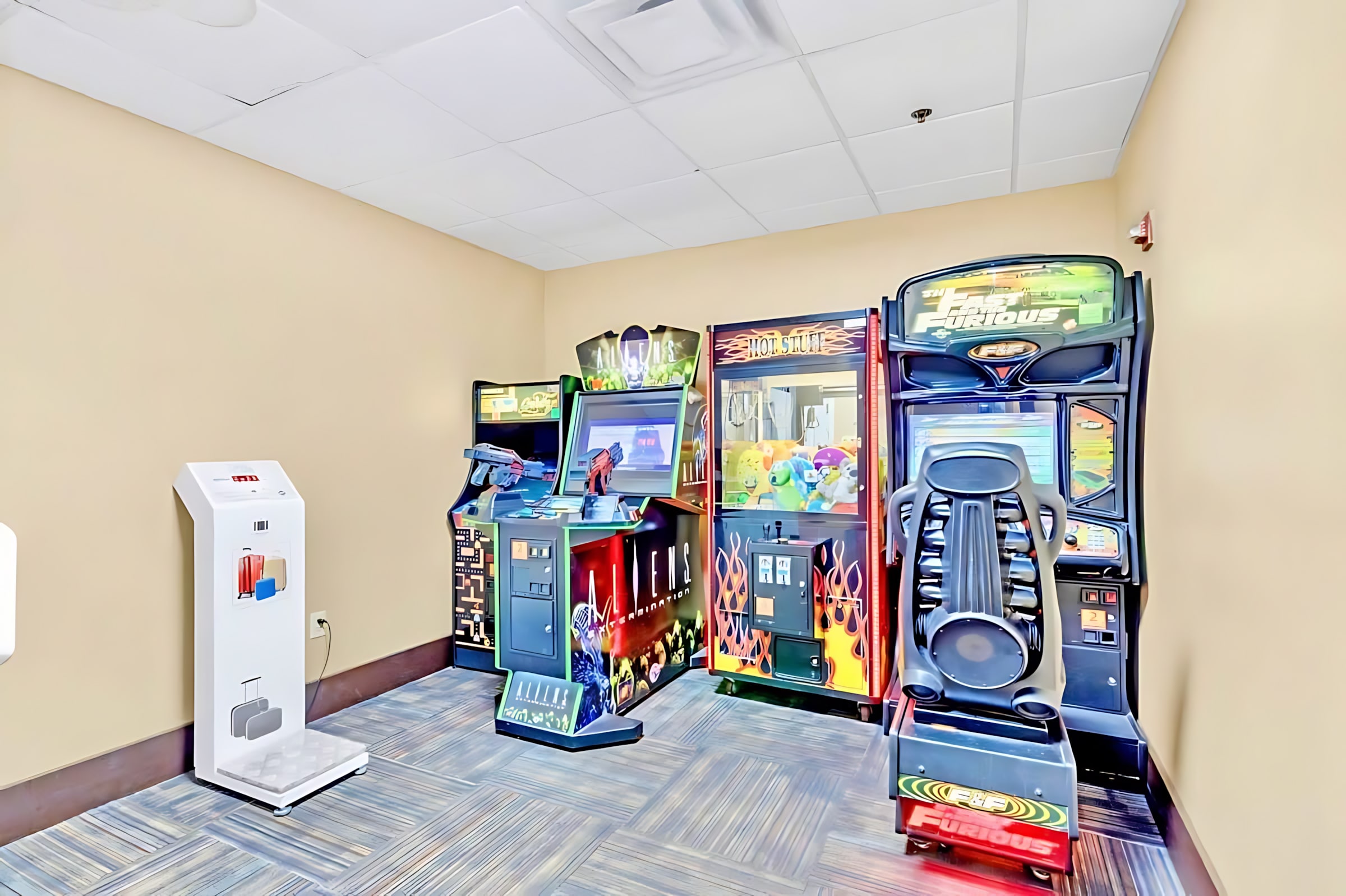 A vibrant game room featuring multiple arcade machines, including racing games and claw machines, set against light beige walls and a patterned carpet. A hand sanitizer dispenser stands near the entrance.