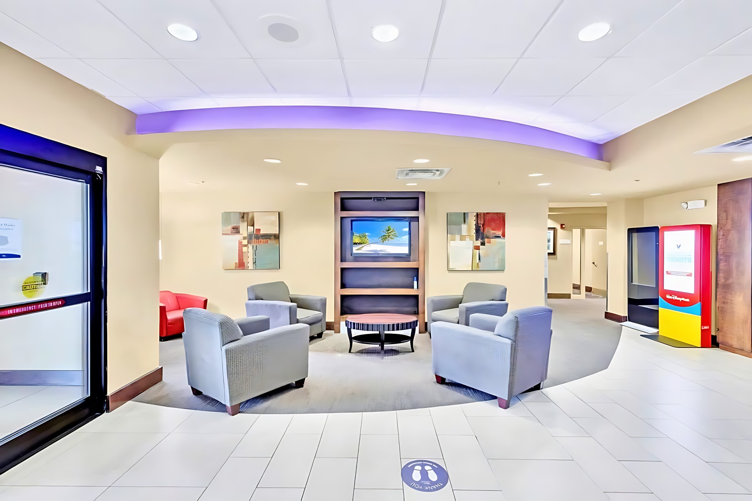 A comfortable communal lounge area with several grey armchairs arranged around a circular coffee table, facing a wall-mounted TV within a dark wood entertainment unit. The space features light beige walls and a patterned carpet.