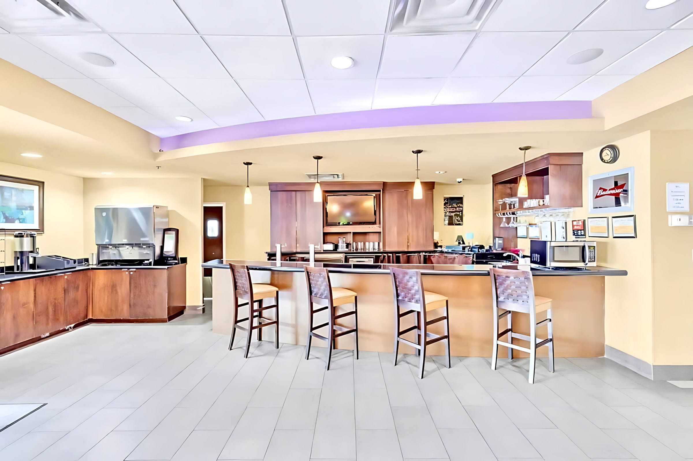 A modern communal bar and serving area with dark wood cabinetry, a long counter with four bar stools, and various appliances like coffee makers, an ice machine, and a microwave.