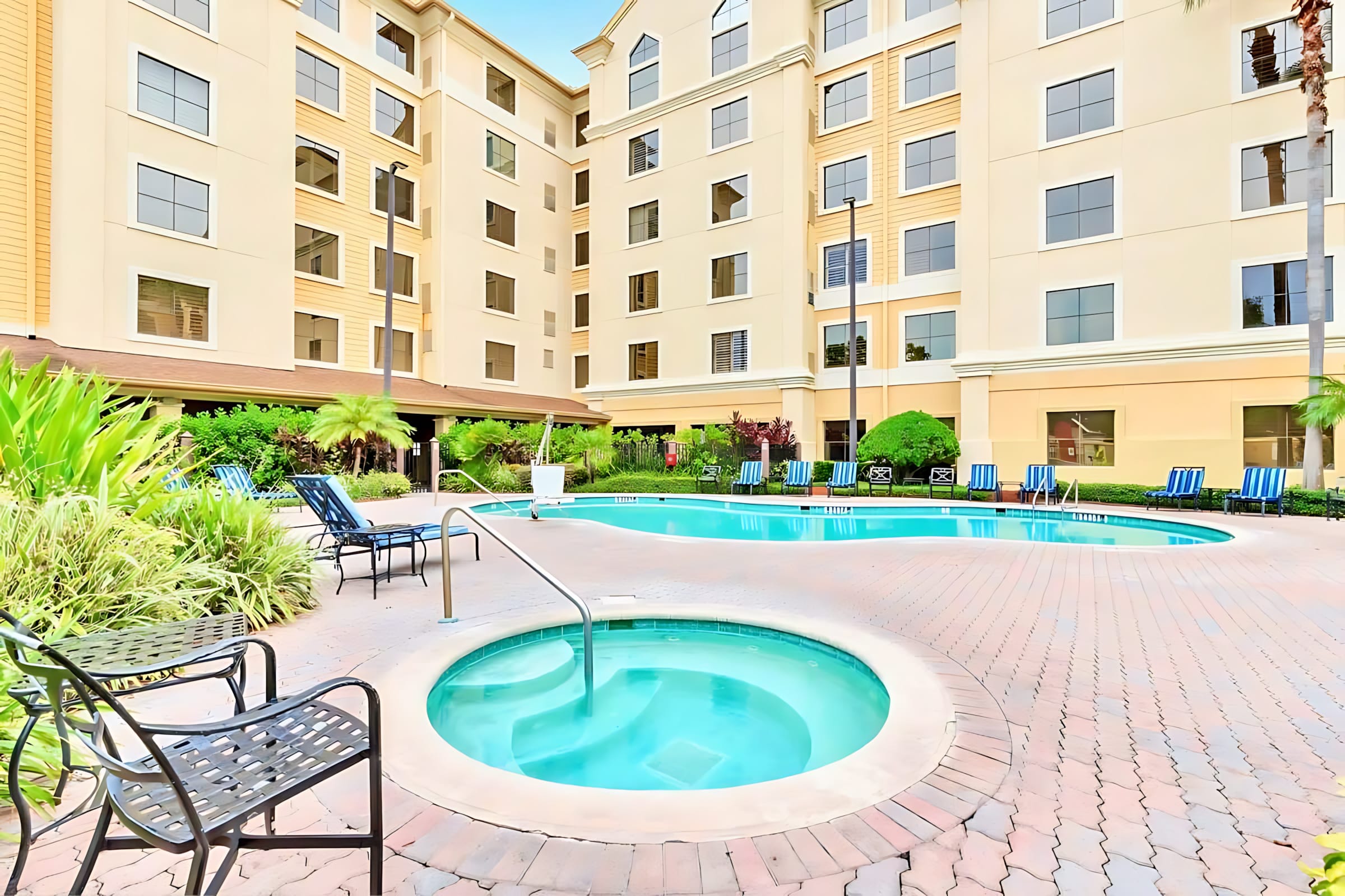 An inviting outdoor pool area featuring a large swimming pool and a separate circular hot tub, surrounded by a patterned brick deck. Lounge chairs and lush tropical landscaping are visible, with the building exterior in the background.