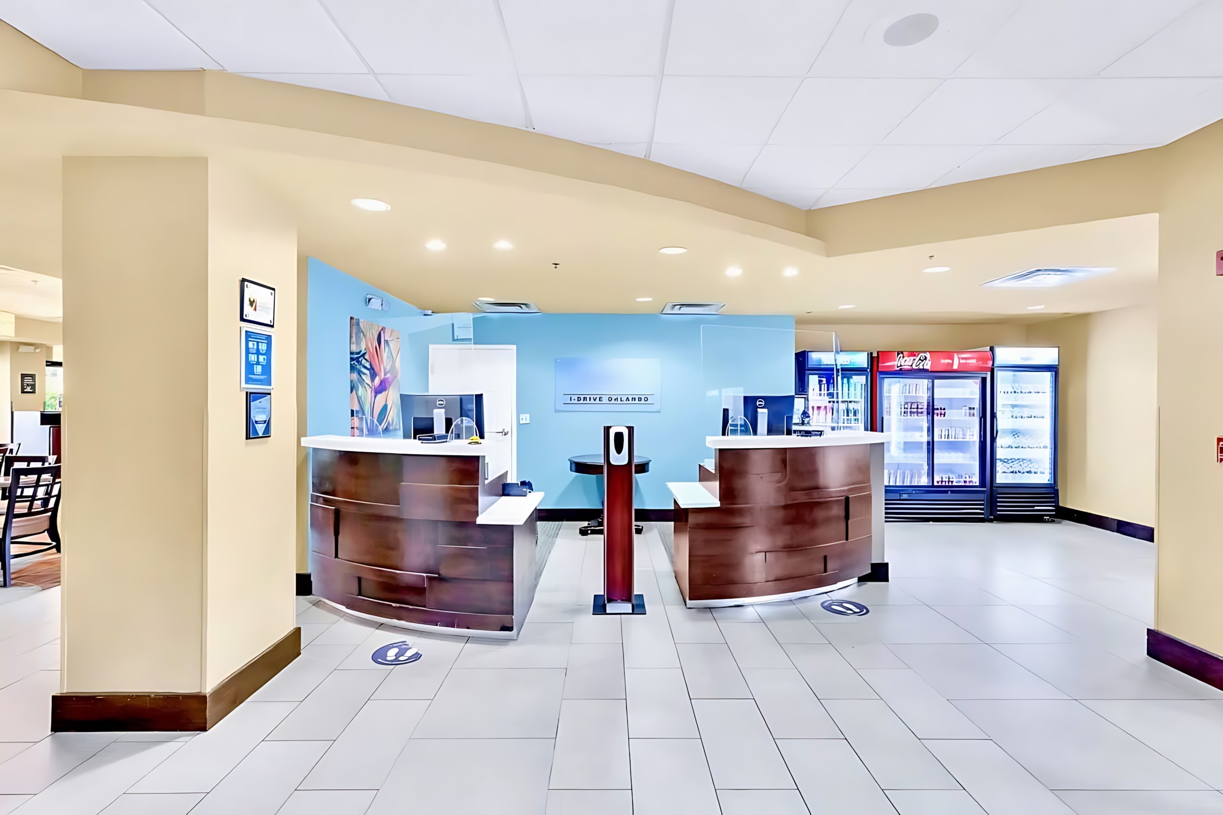 A bright and welcoming reception area featuring two curved dark wood desks with light blue accent walls, a central hand sanitizer station, and refrigerators stocked with beverages.