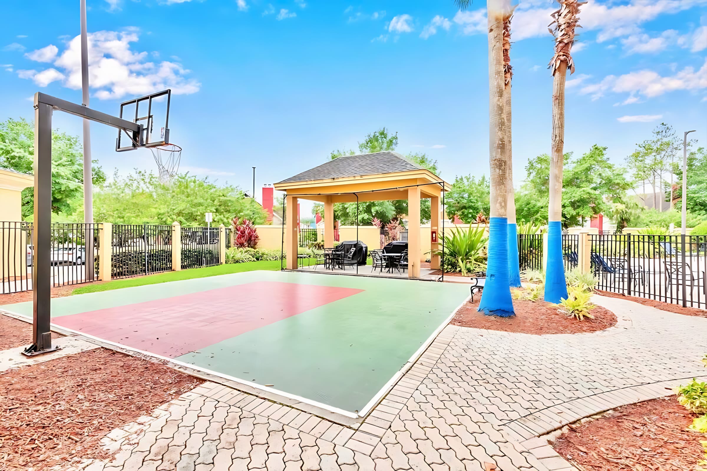 An outdoor recreational area featuring a half-basketball court with green and red sections, a shaded gazebo with seating, and tall palm trees, all enclosed by a black metal fence.