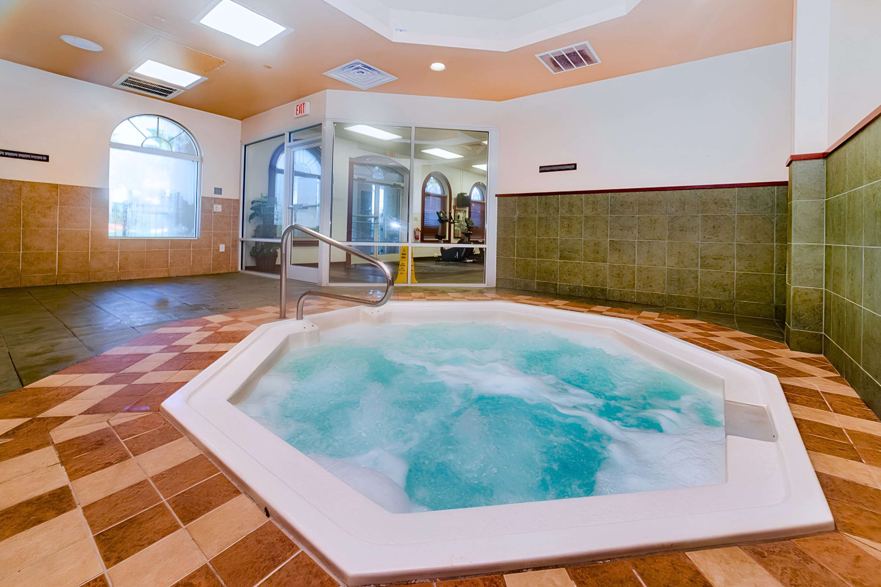 An indoor hot tub with bubbling water is set in a tiled room with warm-toned floor tiles and green and brown wall tiles, featuring large arched windows and a view into a fitness center.