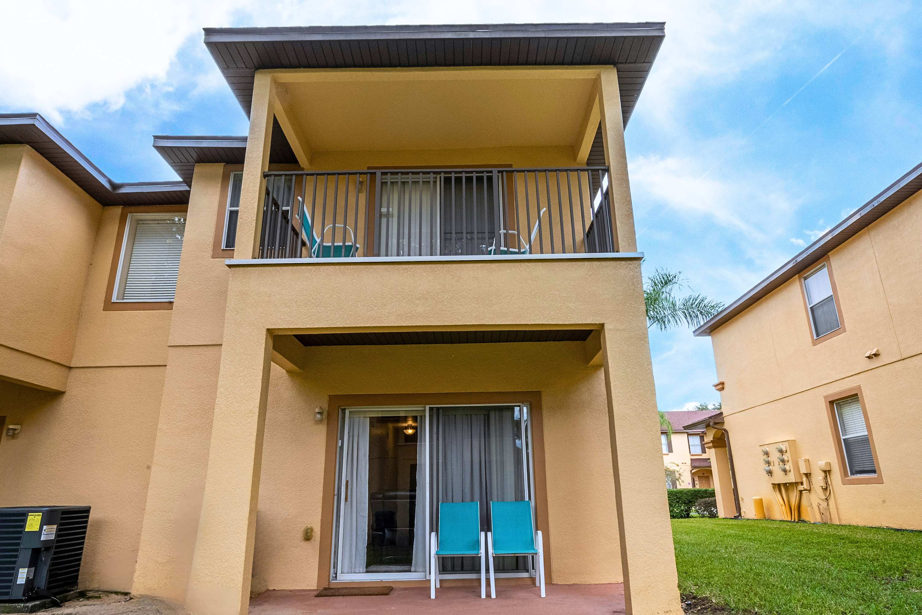 The back of the townhouse features a ground-level patio with sliding glass doors and two teal chairs, topped by a second-story balcony with black railings and additional seating, all against a light yellow stucco facade.