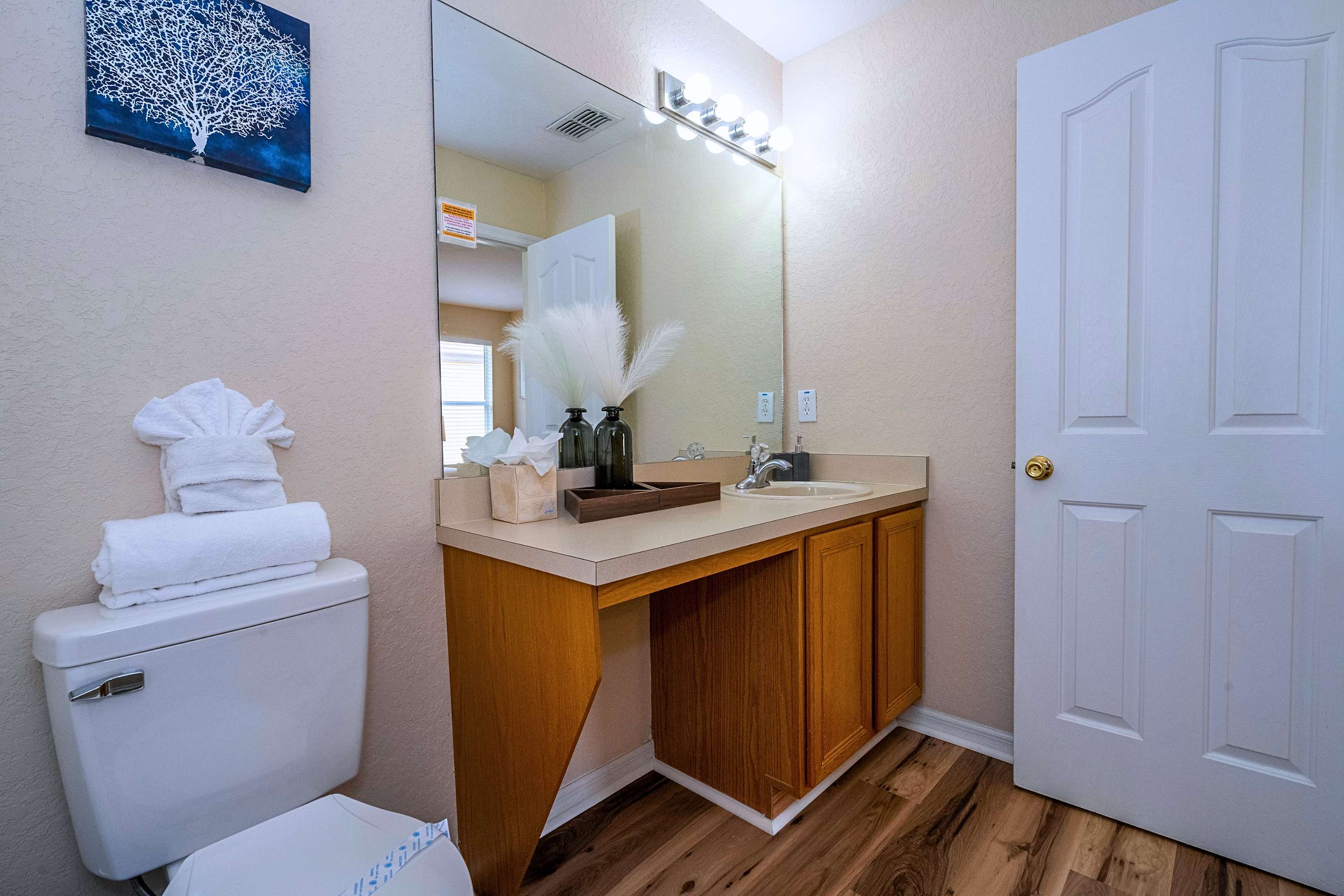 Vanity area of a bathroom with a light wood cabinet, single sink, large mirror, and bright overhead lighting, next to a toilet with folded towels, all on light wood laminate flooring.
