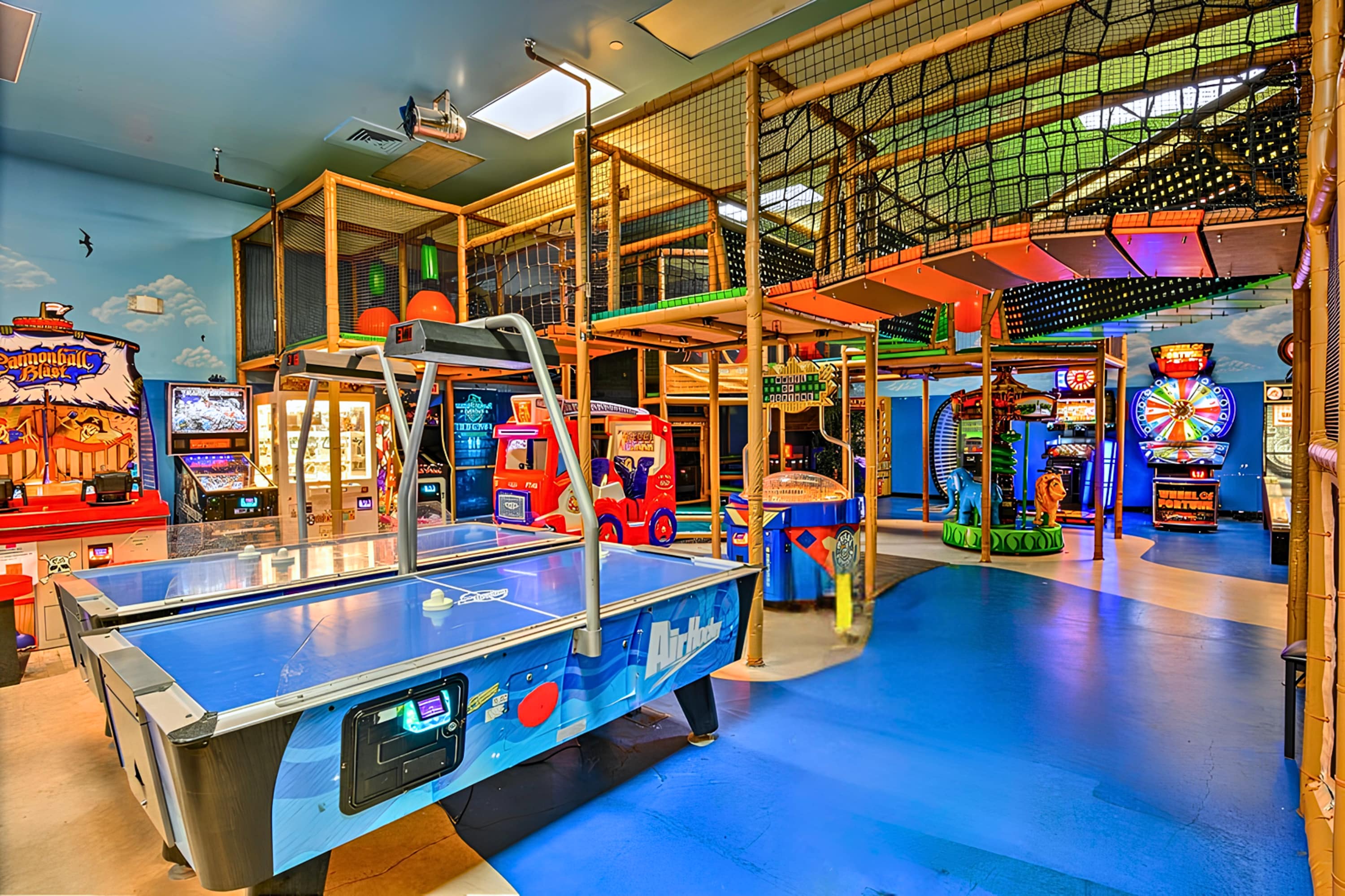 A vibrant game room features multiple arcade machines, an air hockey table, and a large multi-level indoor play structure with nets and slides, all on a bright blue floor.