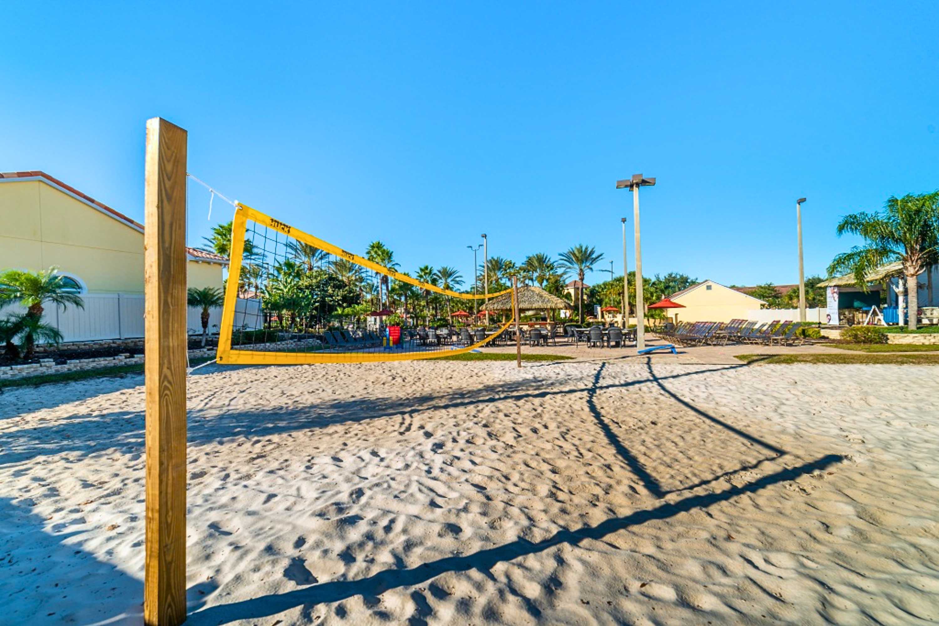 A sunny outdoor sand volleyball court with a yellow net is ready for play, surrounded by green grass, palm trees, and resort buildings under a clear blue sky.