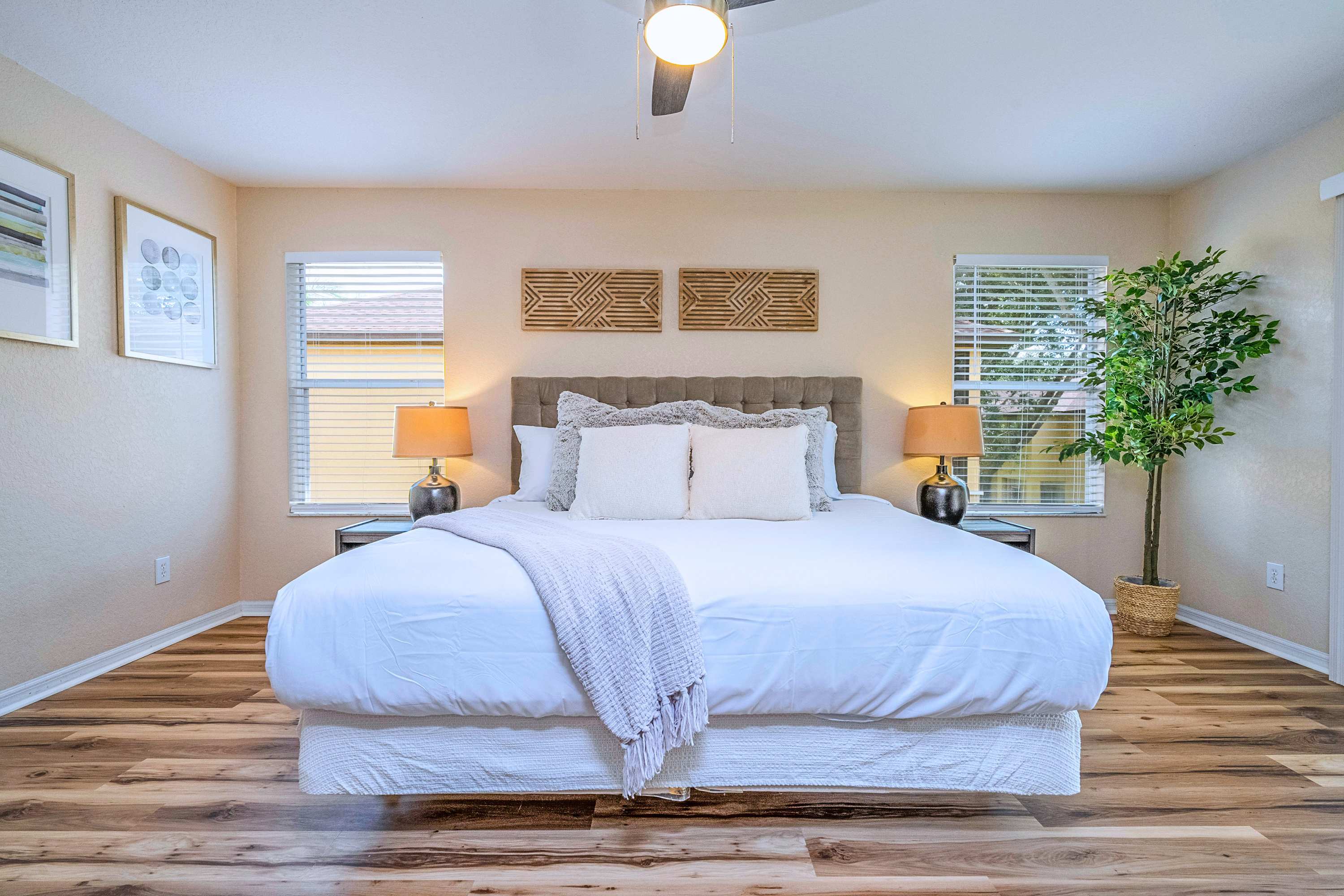 Frontal view of a spacious bedroom with a king-sized bed, white bedding, and a grey upholstered headboard, flanked by two nightstands with lamps, and two windows with blinds, featuring decorative wall art.