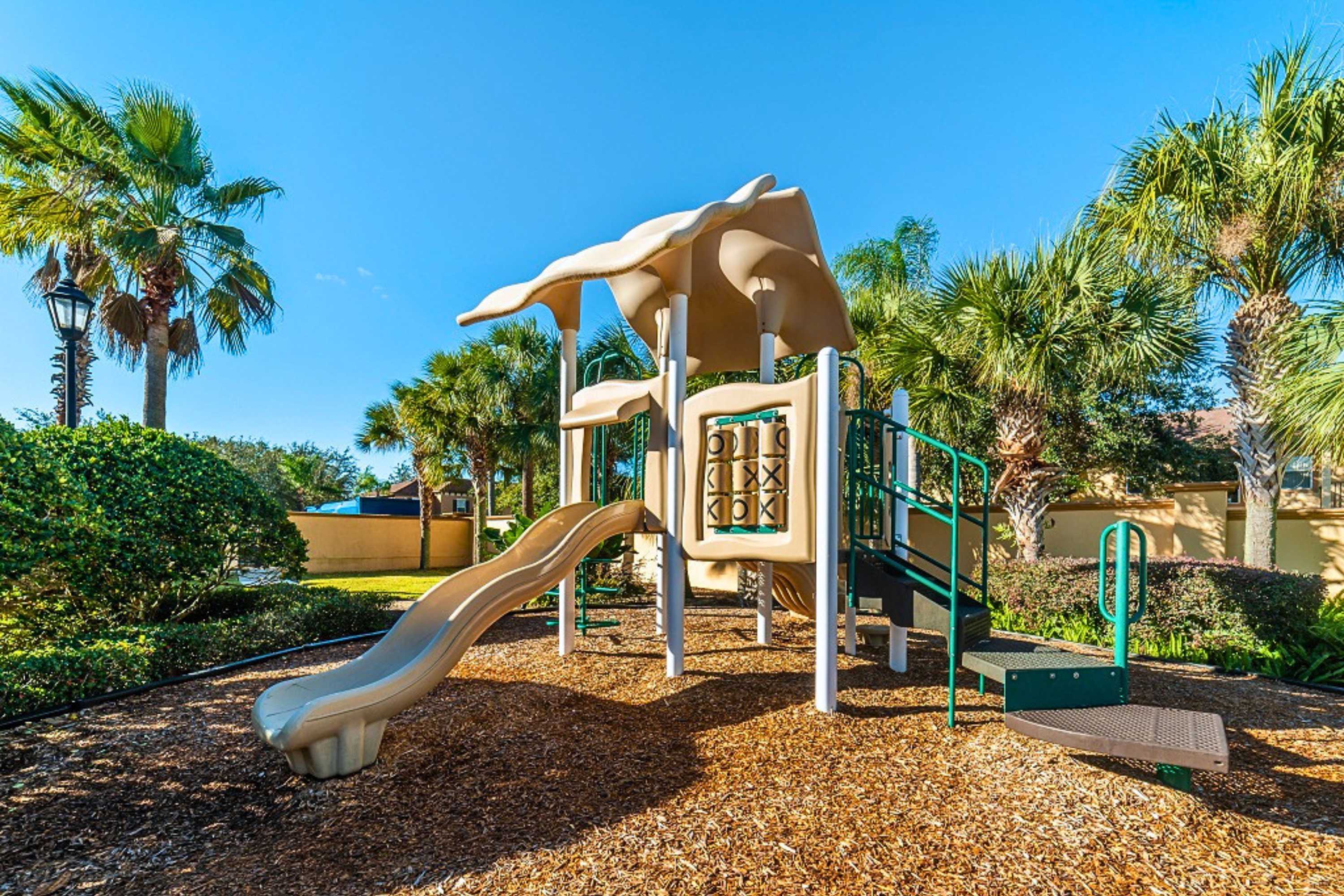 A colorful outdoor playground features a slide, climbing structures, and a tic-tac-toe panel, set on a wood chip surface and surrounded by green bushes and palm trees.