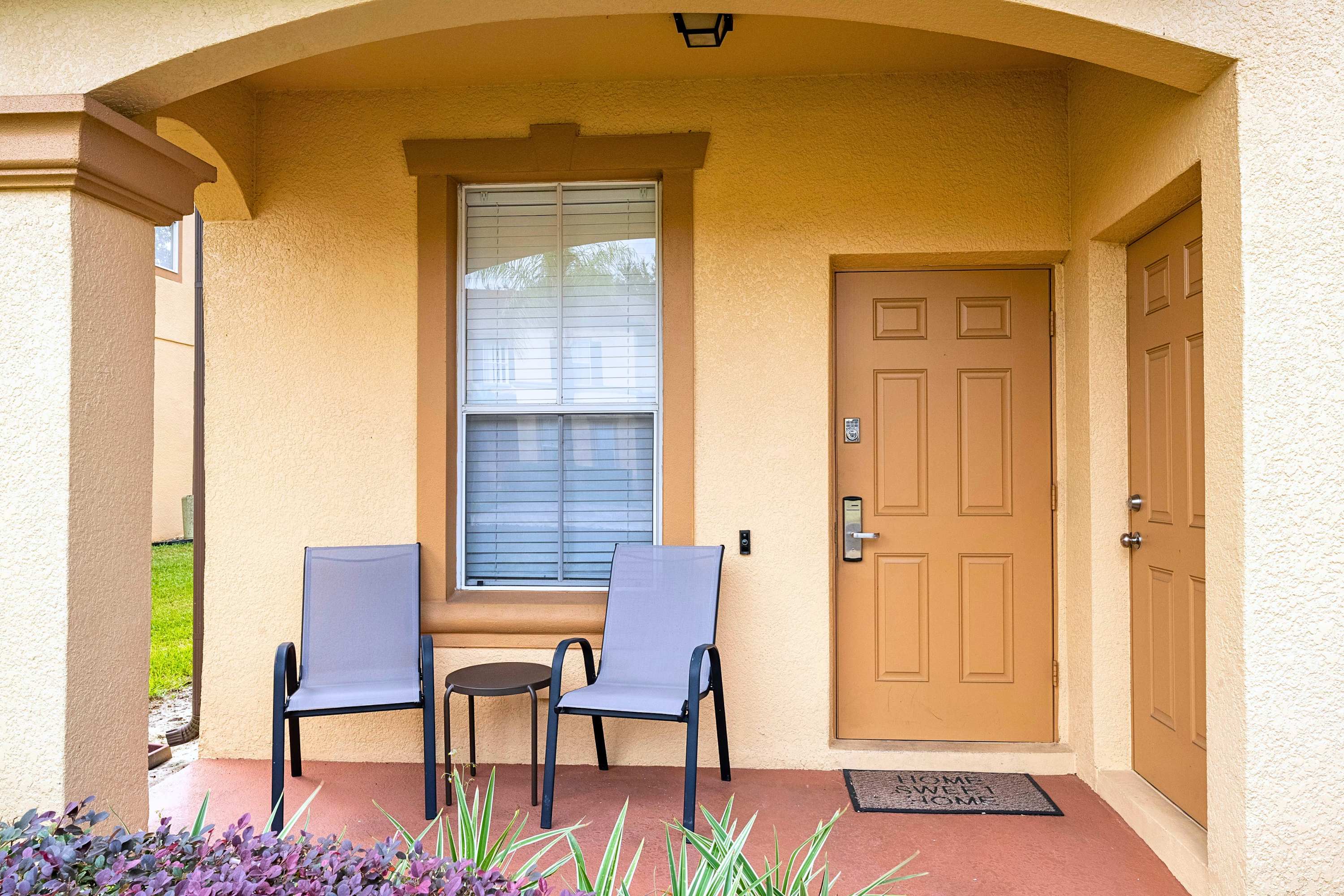 The front porch features a light brown door with a 'HOME SWEET HOME' mat, a large window with blinds, and two grey outdoor chairs with a small table, all under a covered archway.