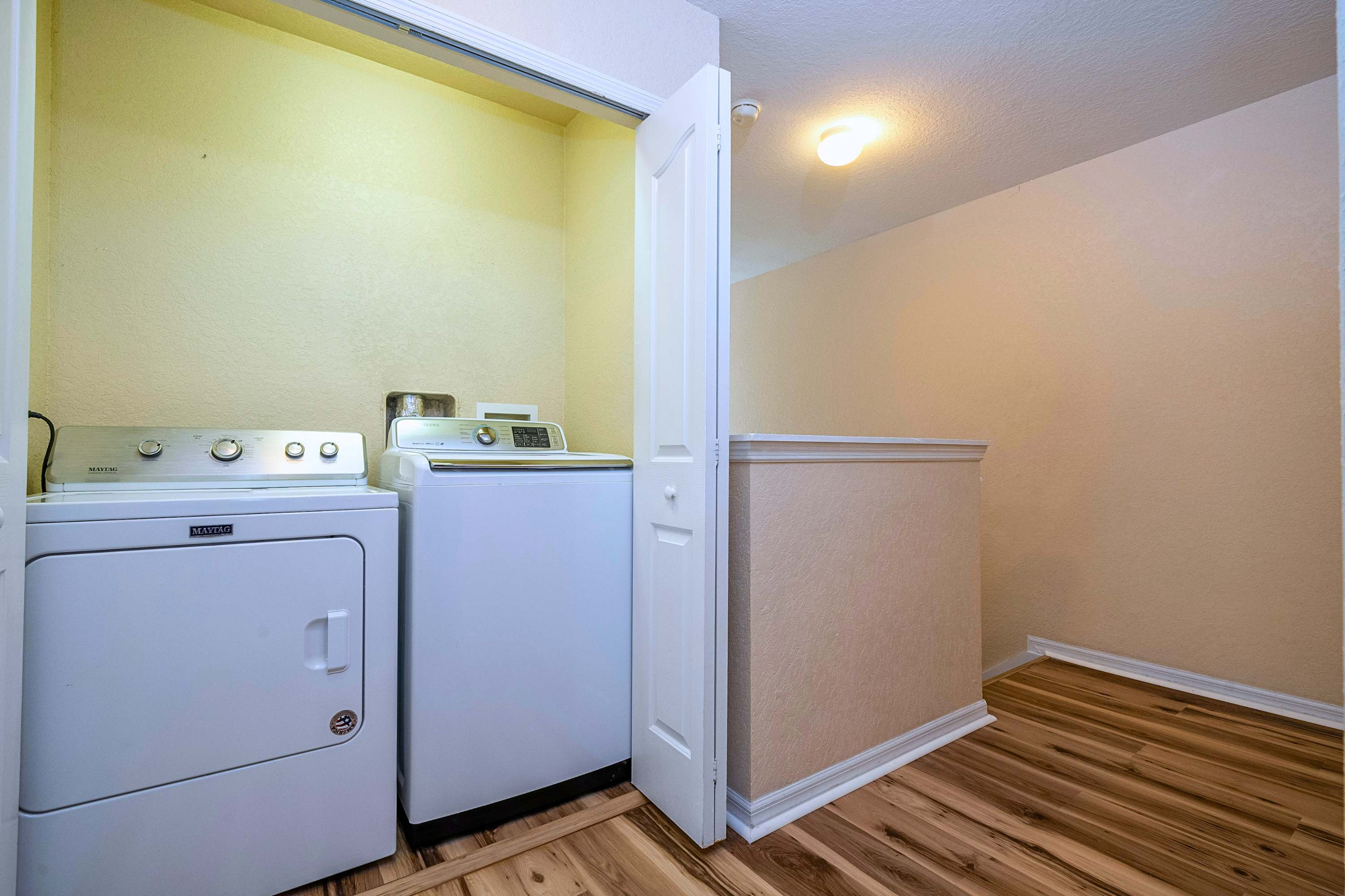 A compact laundry area features a white Maytag washer and dryer set, neatly tucked into an alcove with light yellow walls and light wood-look flooring.