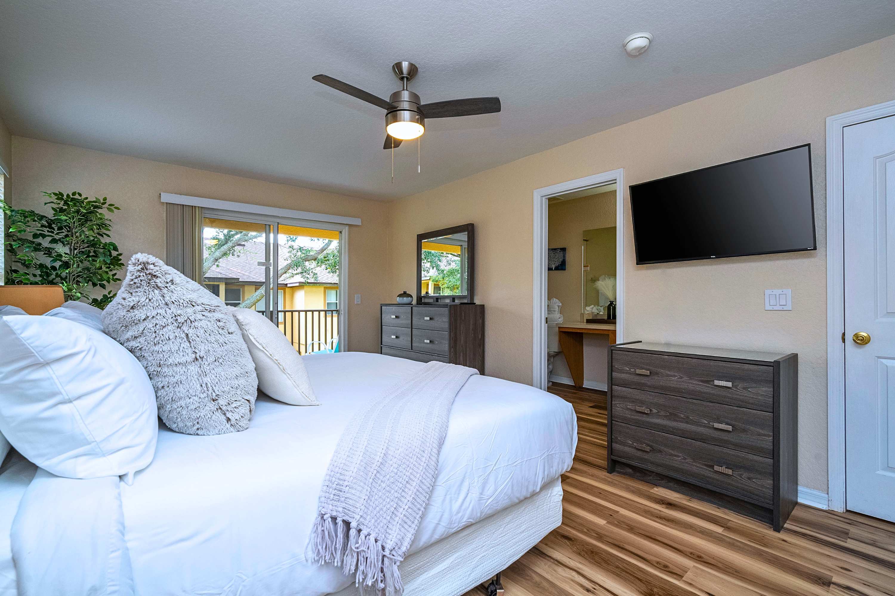 Master bedroom with a large bed featuring white bedding and a grey textured headboard, a wall-mounted TV, and a dark grey dresser with a mirror, with access to a balcony through sliding glass doors.