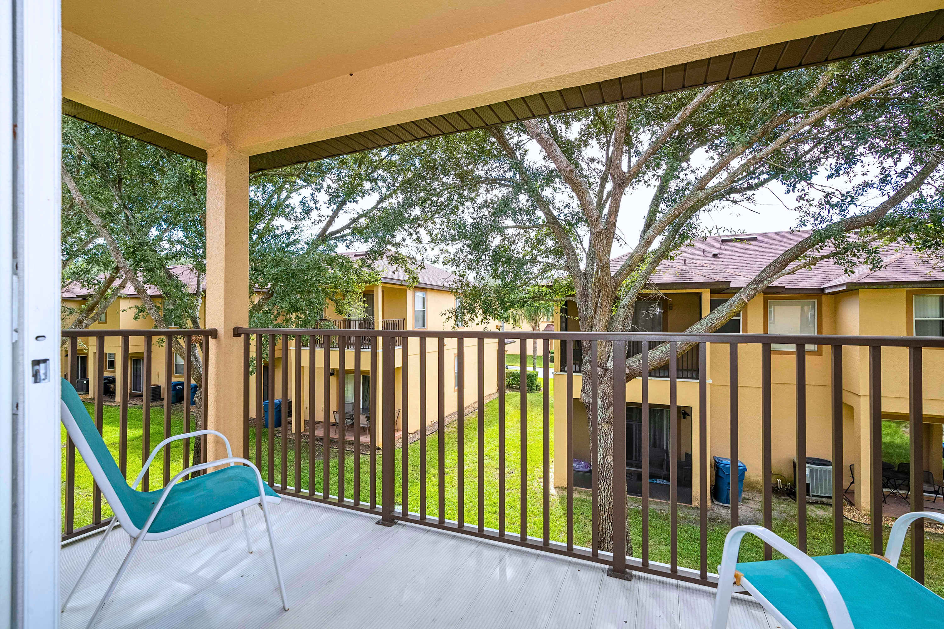 Close-up view of the private balcony with a metal railing, featuring two teal outdoor chairs and a scenic view of mature trees and the surrounding townhome complex.