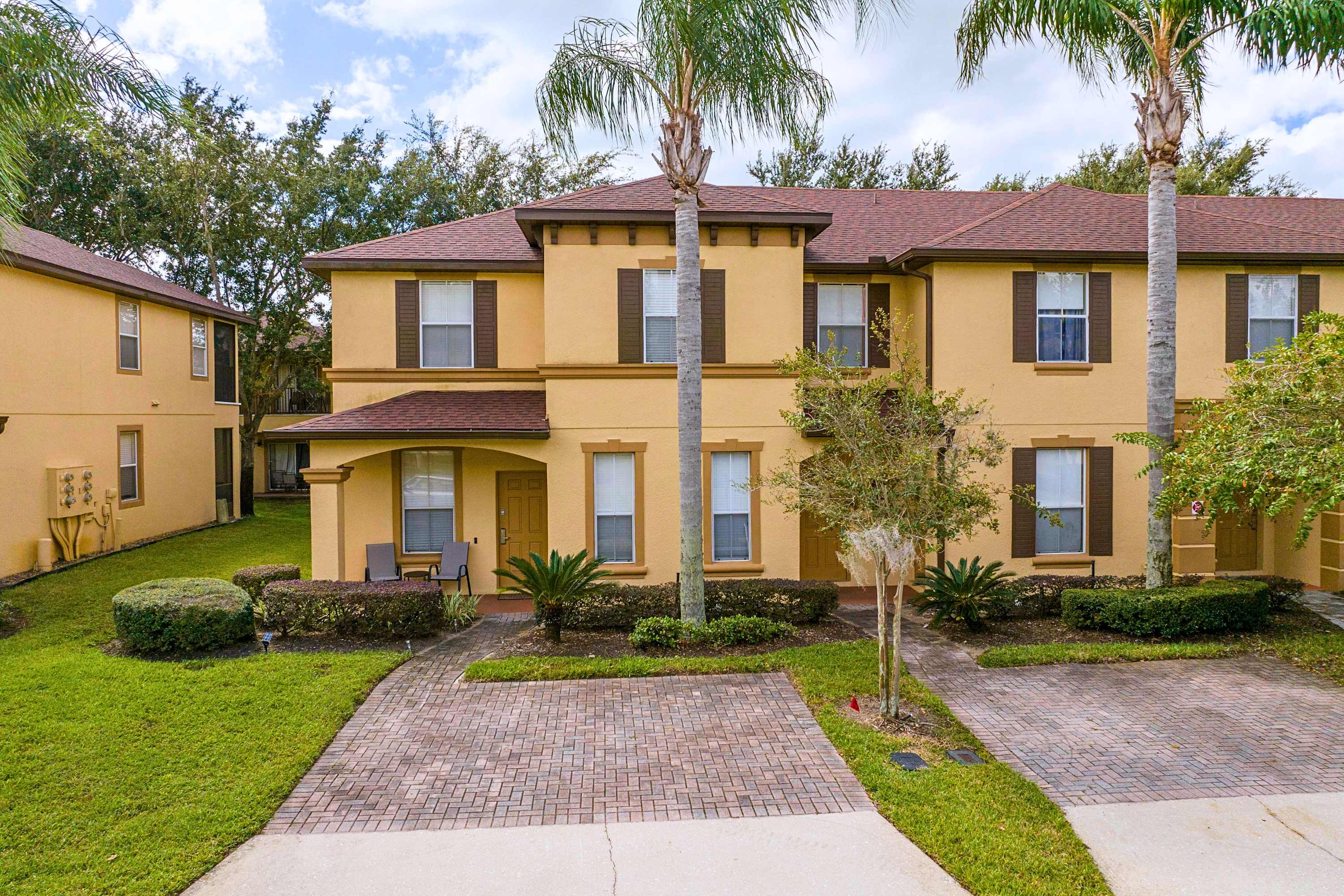An inviting multi-unit townhouse building with warm yellow stucco and dark brown shutters is surrounded by lush green landscaping and tall palm trees, featuring a brick-paved driveway.