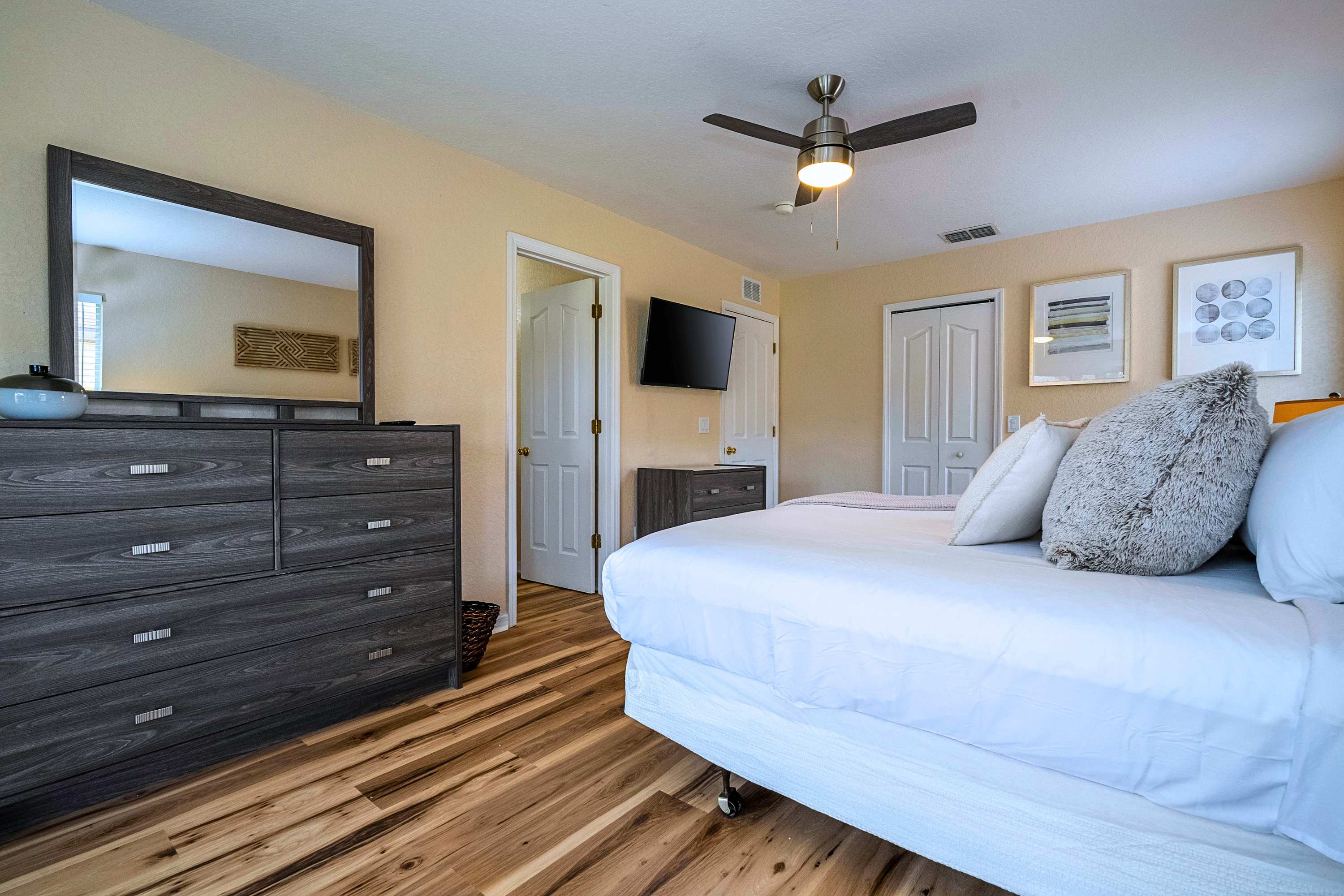 Angled view of the master bedroom showing a large bed with white bedding, a dark grey dresser with a mirror, a wall-mounted TV, and two white closet doors, all on light wood laminate flooring.