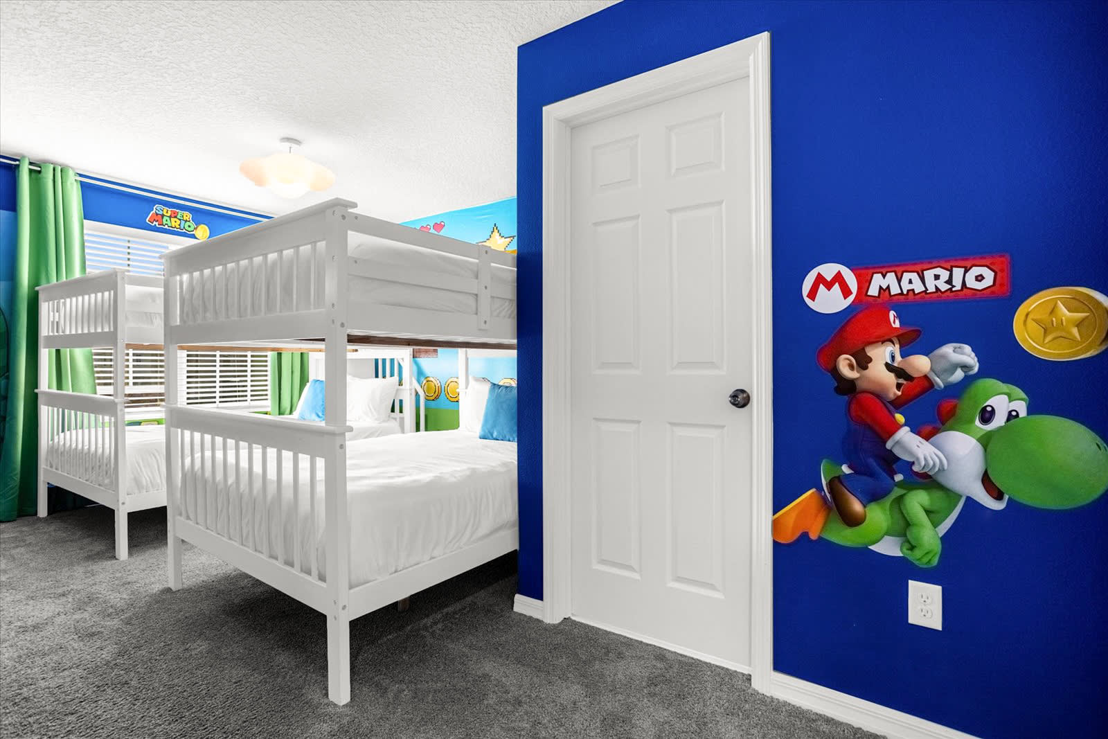 A Super Mario-themed bedroom featuring two white wooden bunk beds with white linens and blue accent pillows, set against a vibrant blue wall with a large Mario and Yoshi decal. A white door and light gray carpet complete the playful space.