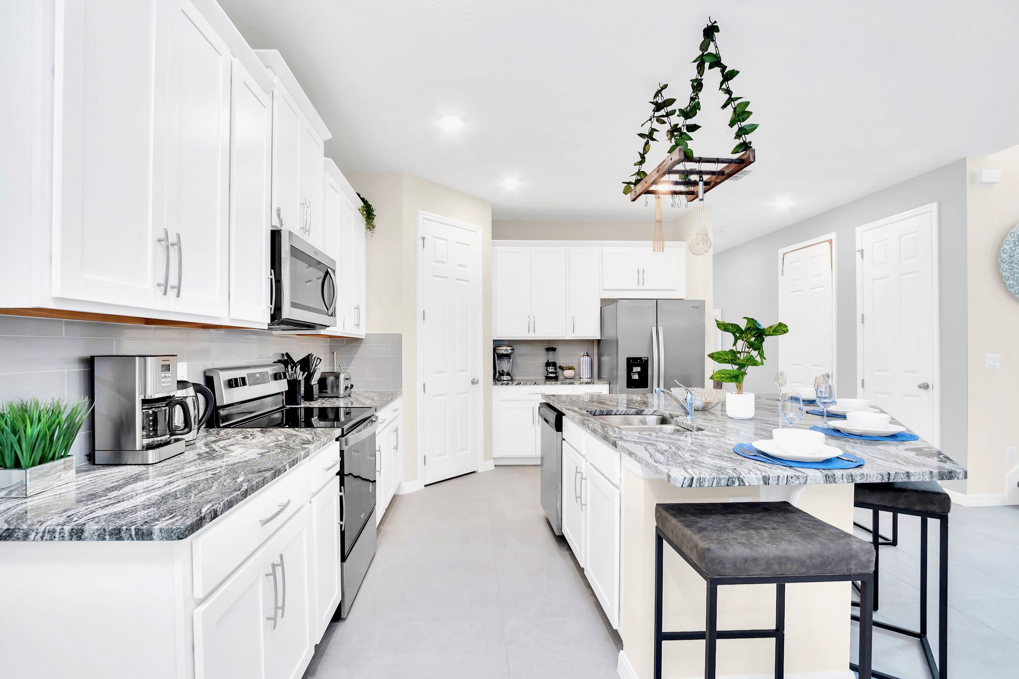 A contemporary kitchen features white shaker cabinets, speckled granite countertops, and stainless steel appliances including a range, microwave, and refrigerator. A large island with a sink and dishwasher provides additional counter space and seating.