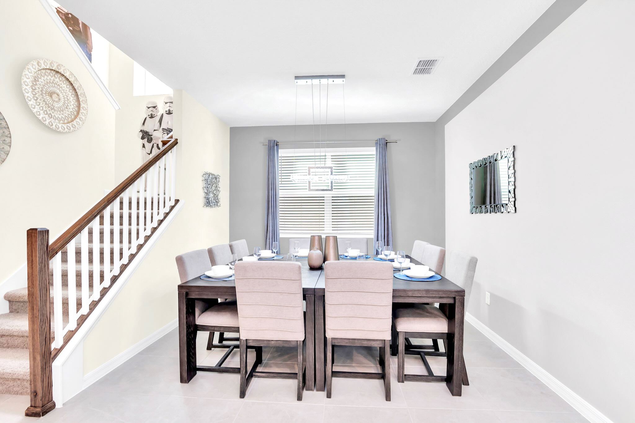 A secondary dining area features a dark wood table with eight upholstered chairs, positioned beneath a modern linear chandelier. A carpeted staircase with white railings and Star Wars decor is visible to the left.