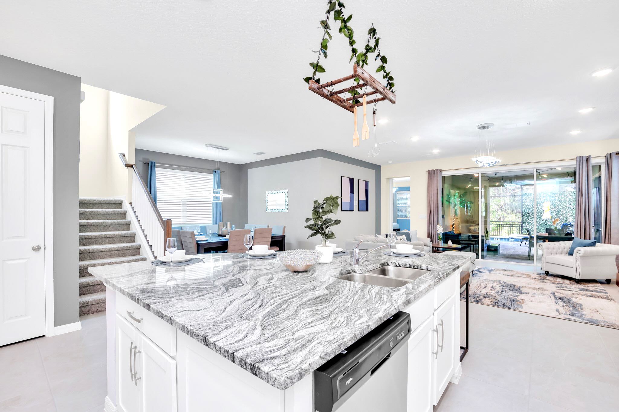 A modern kitchen island with a granite countertop and double sink serves as a breakfast bar, looking towards a formal dining area and a carpeted staircase. White cabinetry and light tile flooring complete the bright, open space.