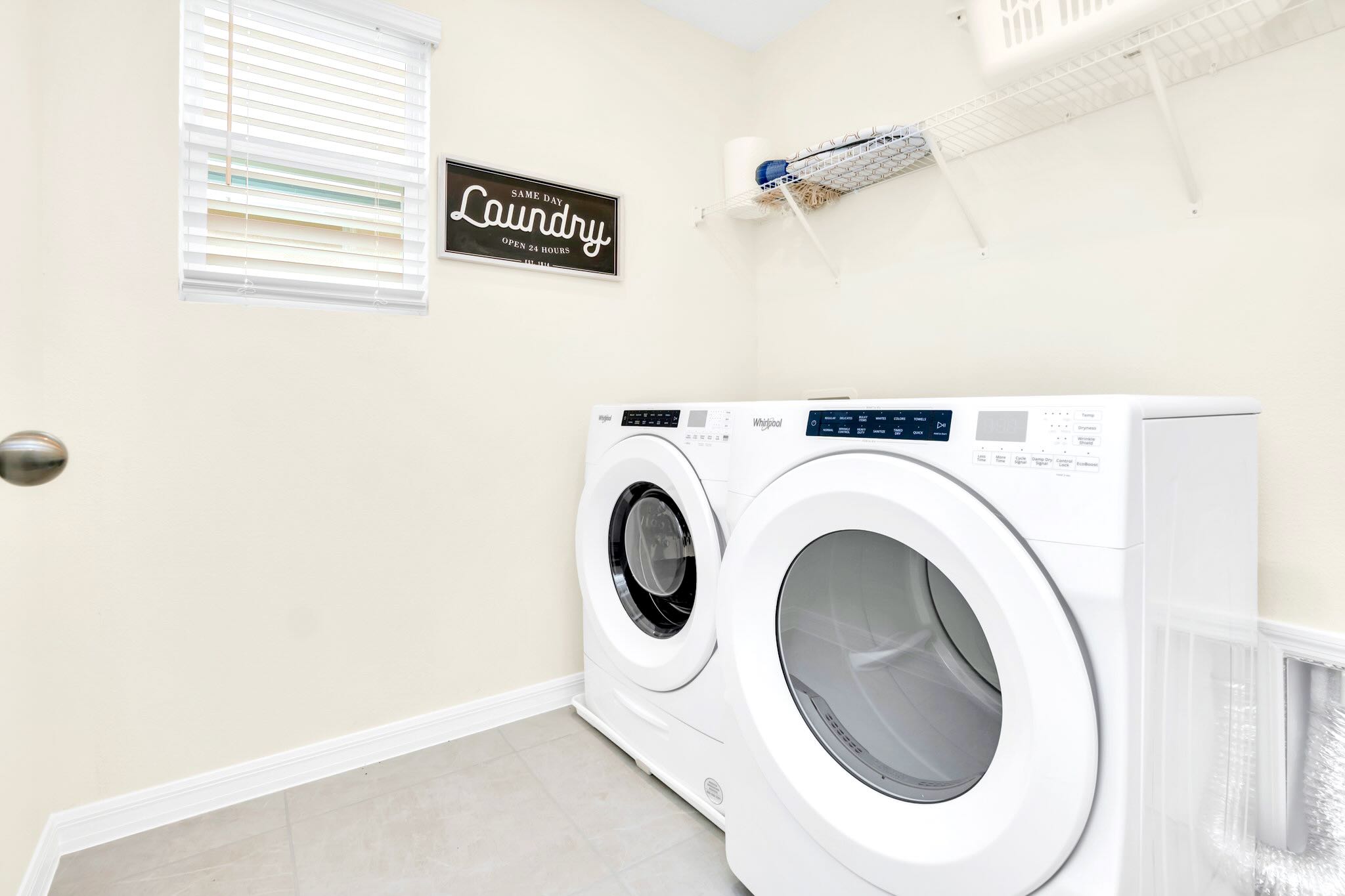 A functional laundry room equipped with a white front-loading washer and dryer set, a window with white blinds, and a wall-mounted shelf for storage. A decorative sign that reads "SAME DAY LAUNDRY OPEN 24 HOURS" hangs on the wall.